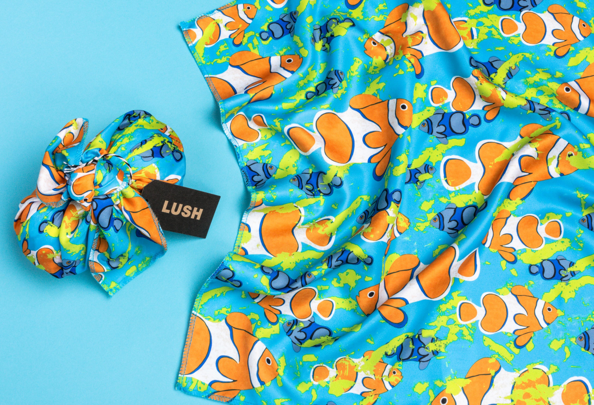 A turqouise blue, Clown Fish Knot Wrap, is laid out flat, with another used to wrap two bath bombs, complete with a gift tag.