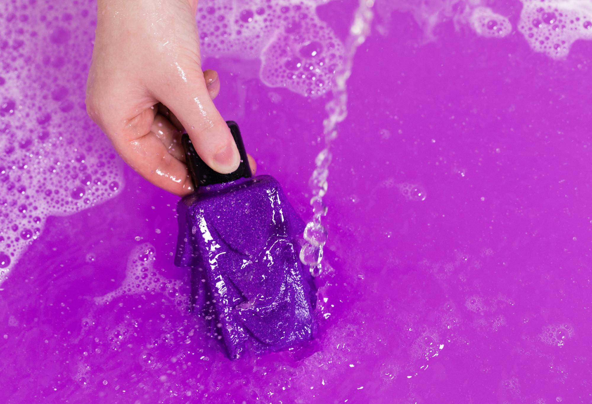 Purple Cocktail is being held by its black wax 'lid' under a gentle stream of water, and in bright purple water, adding bubbles.
