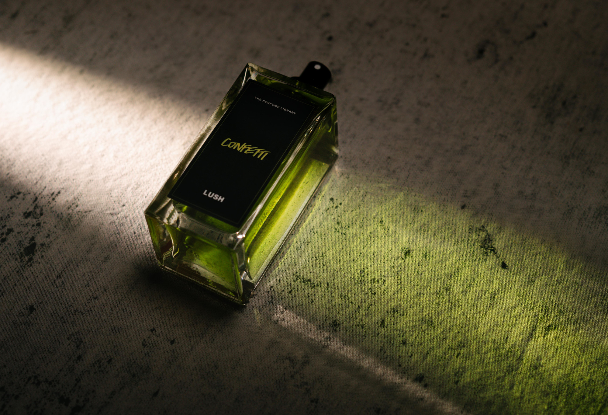 Confetti, a bottled, black label perfume, lies flat, with light shining through it, creating a green cast of light.