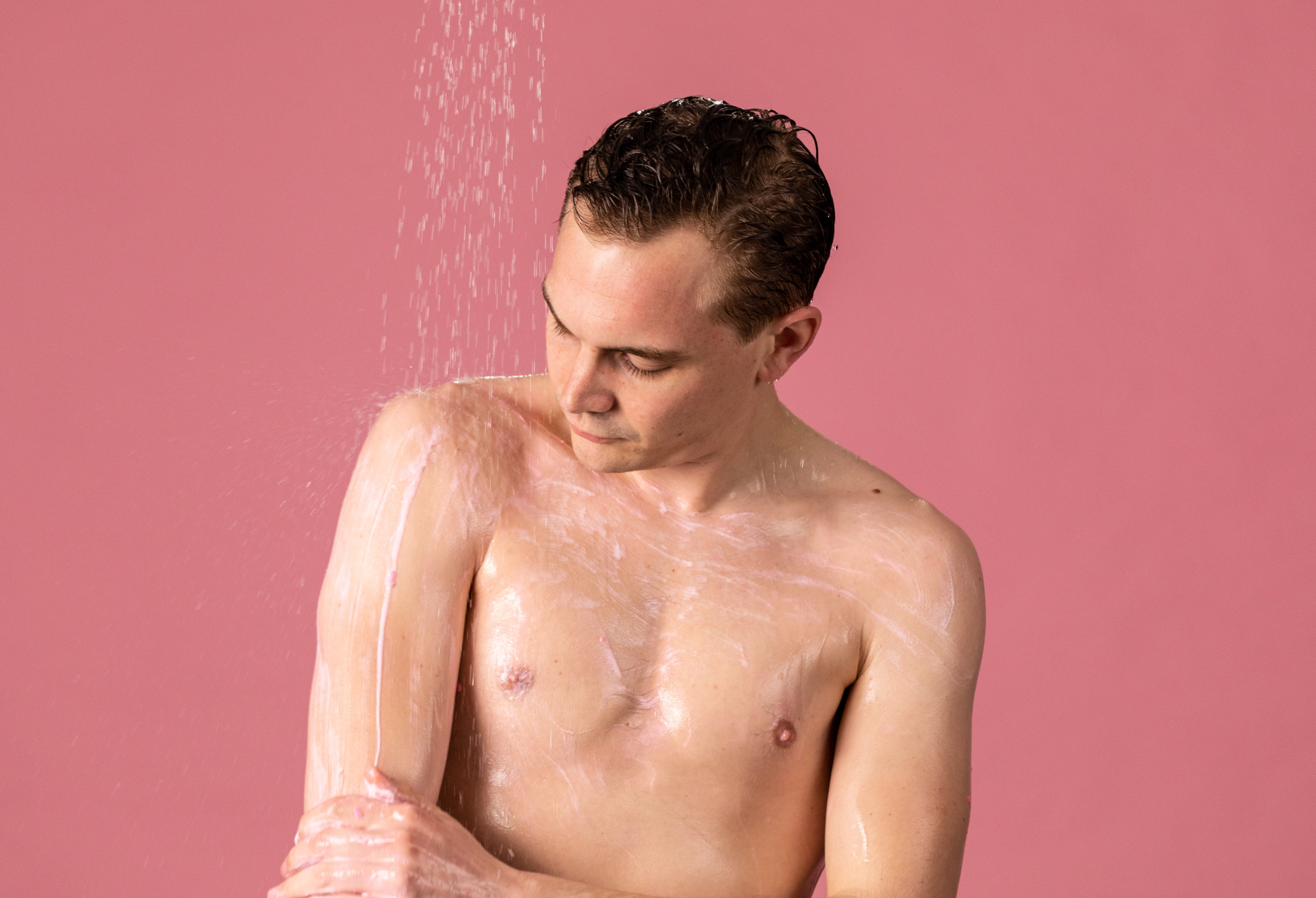 A person, whose upper body is covered in delicate, light pink foam, stands under a shower looking pensive as they rub their arm.