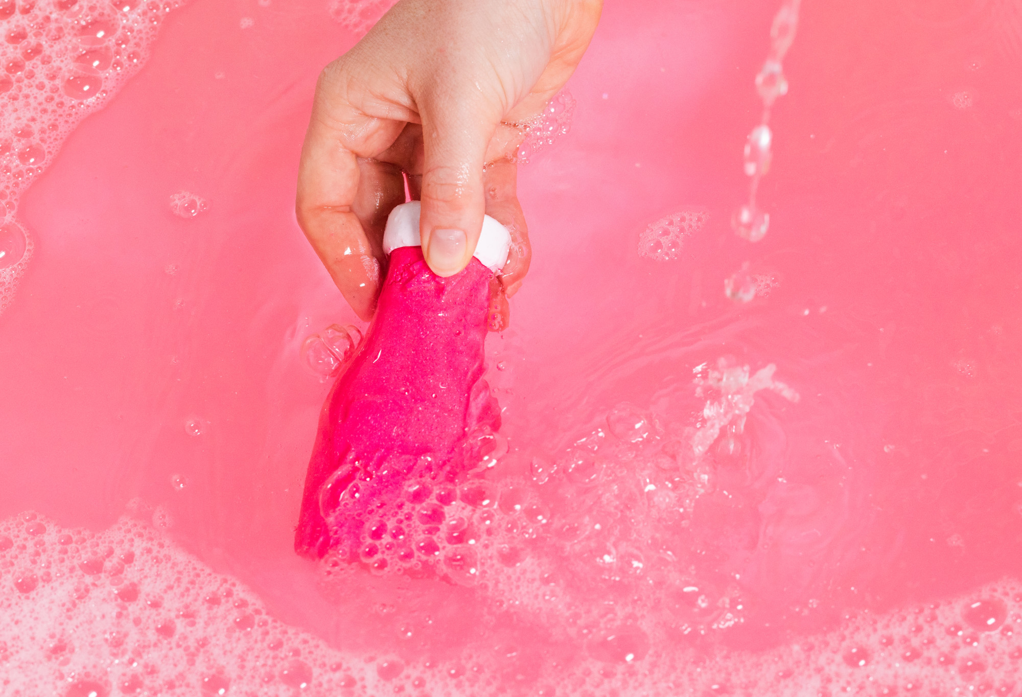 Pink Dirty being held near its white wax 'lid' and run through candy pink water, with bubbles trailing behind.