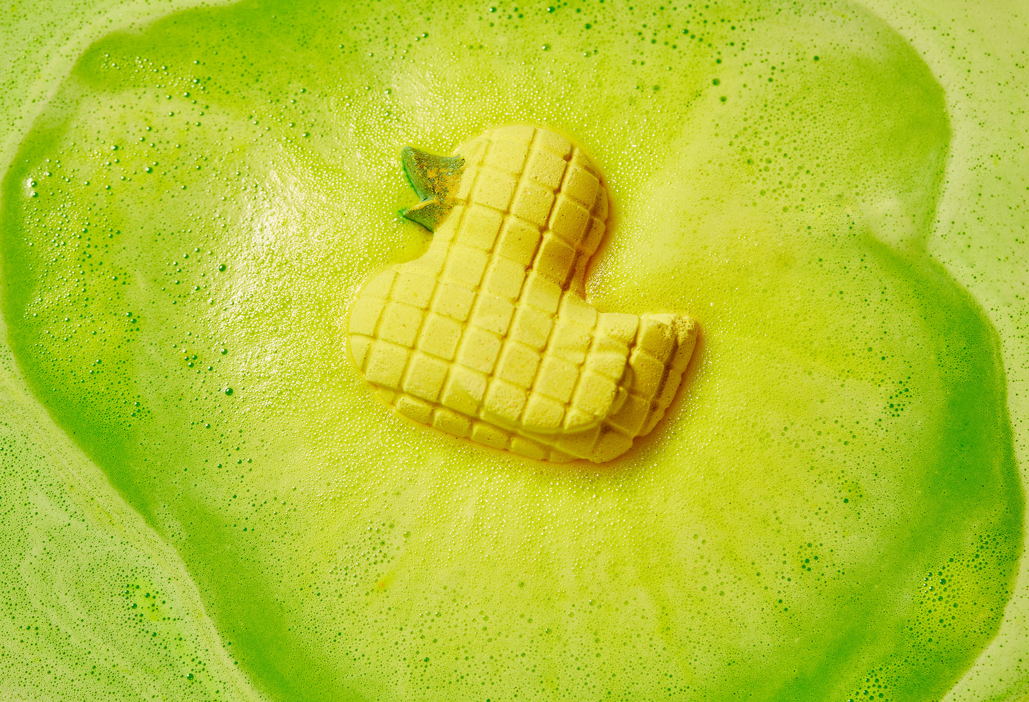 The bath bomb floats on its side in lime green water, giving off yellow hued fizzing foam.