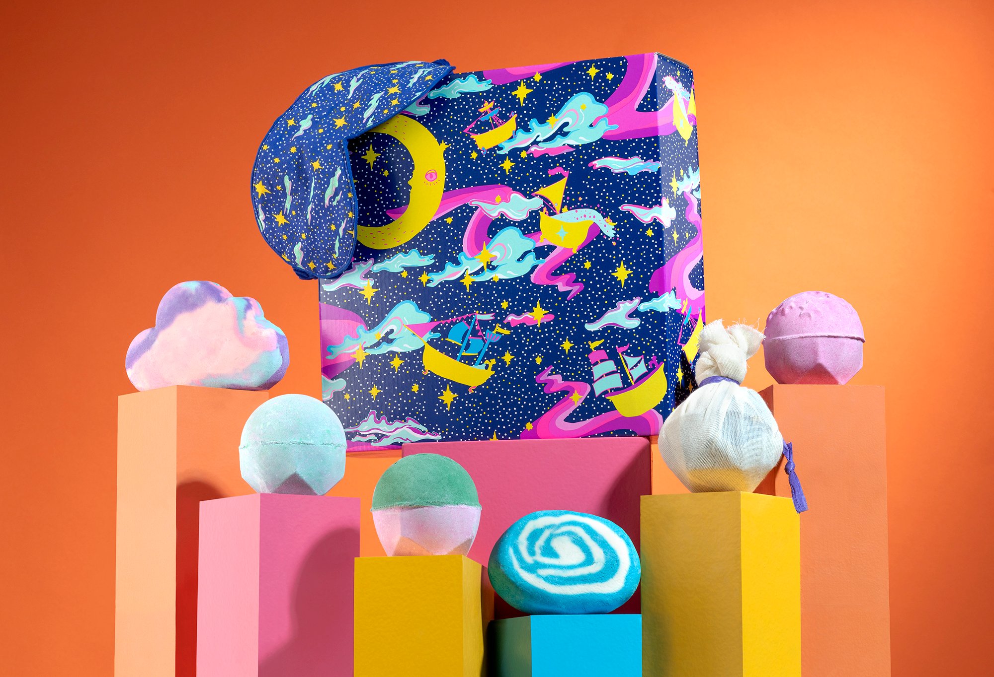 Dreamland gift, in front of an orange background, surrounded by its products on pink, orange, yellow and blue plinths.