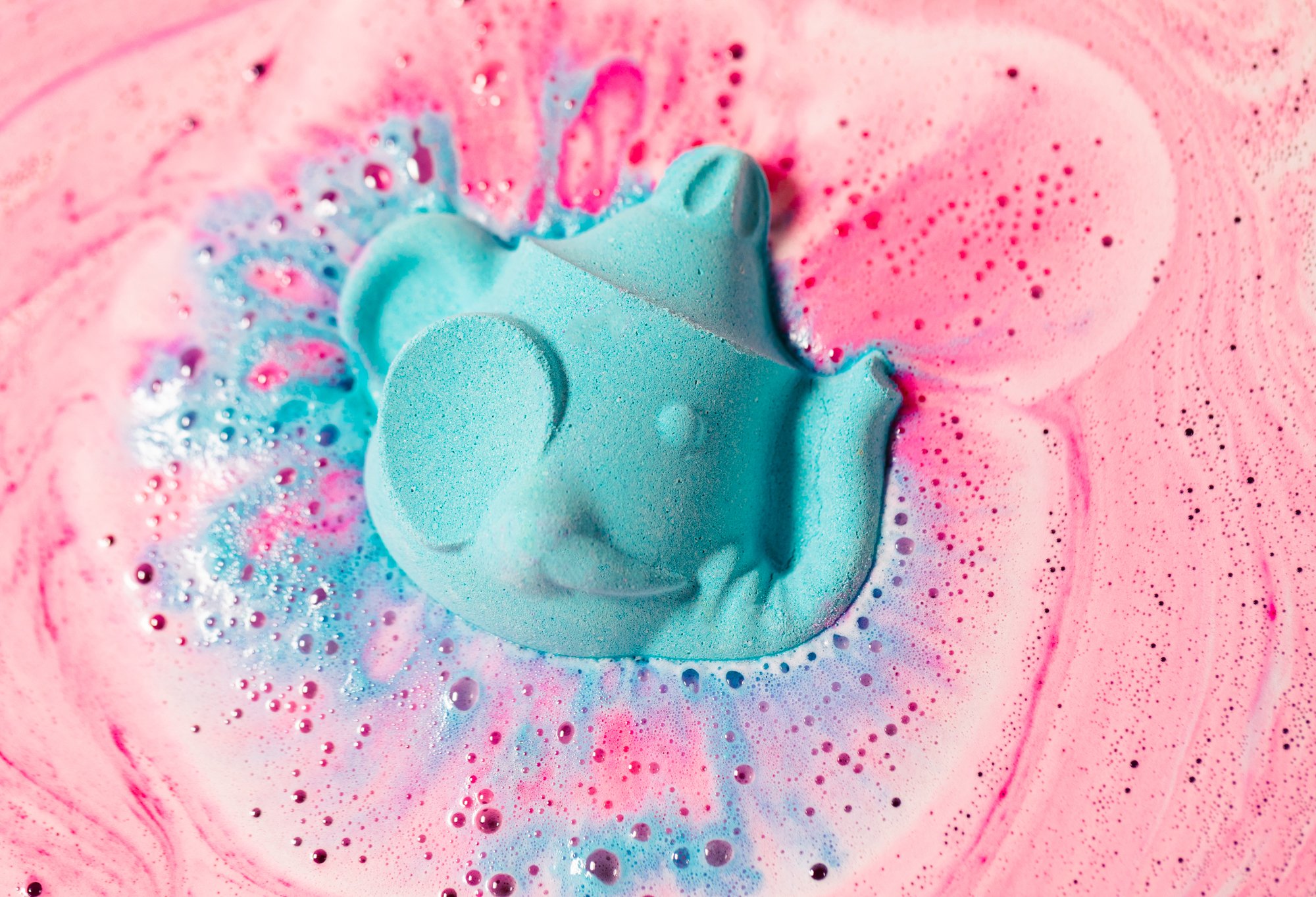 The teapot shaped bath bomb floats in gentle pink foam, its blue half facing up and fizzing out into the water.