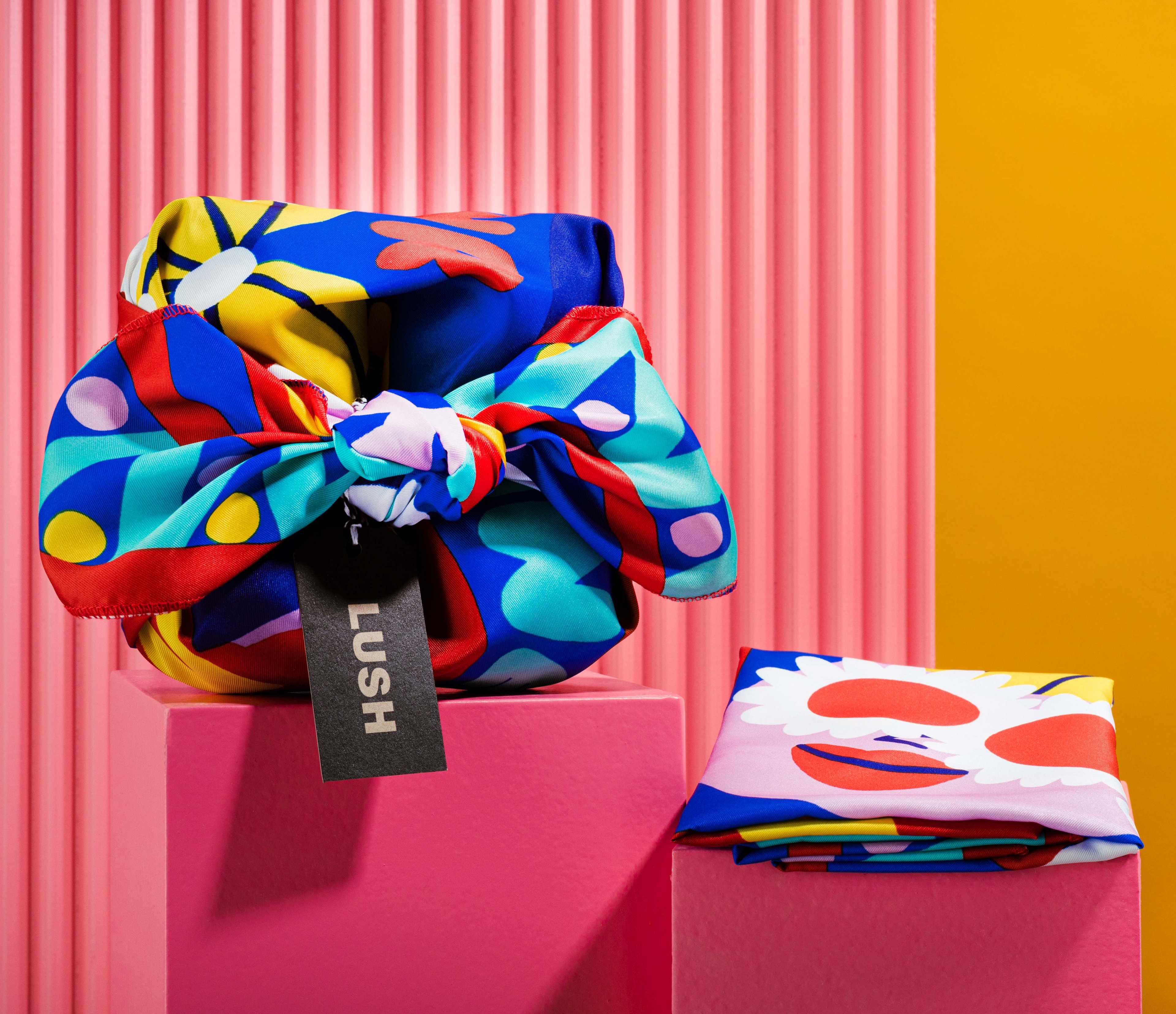 The Knot Wrap is decoratively wrapped with a Lush gift tag, alongside one folded, in front of a pink and yellow background. 