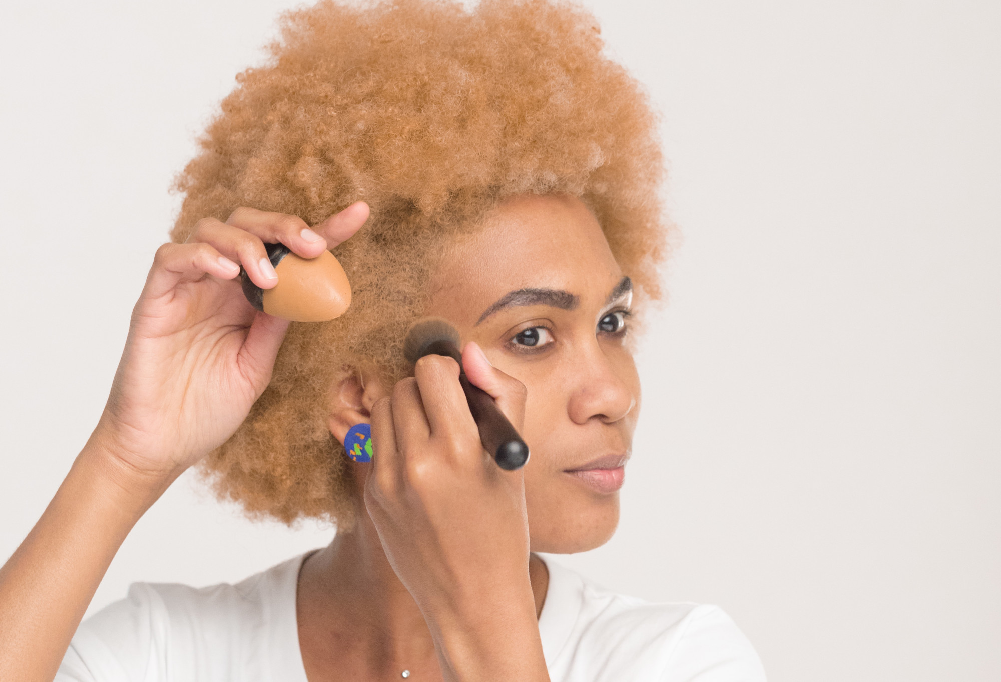 A person with blonde afro hair uses a BFF make up brush to apply a medium dark-warm, egg-shaped solid foundation Slap Stick.