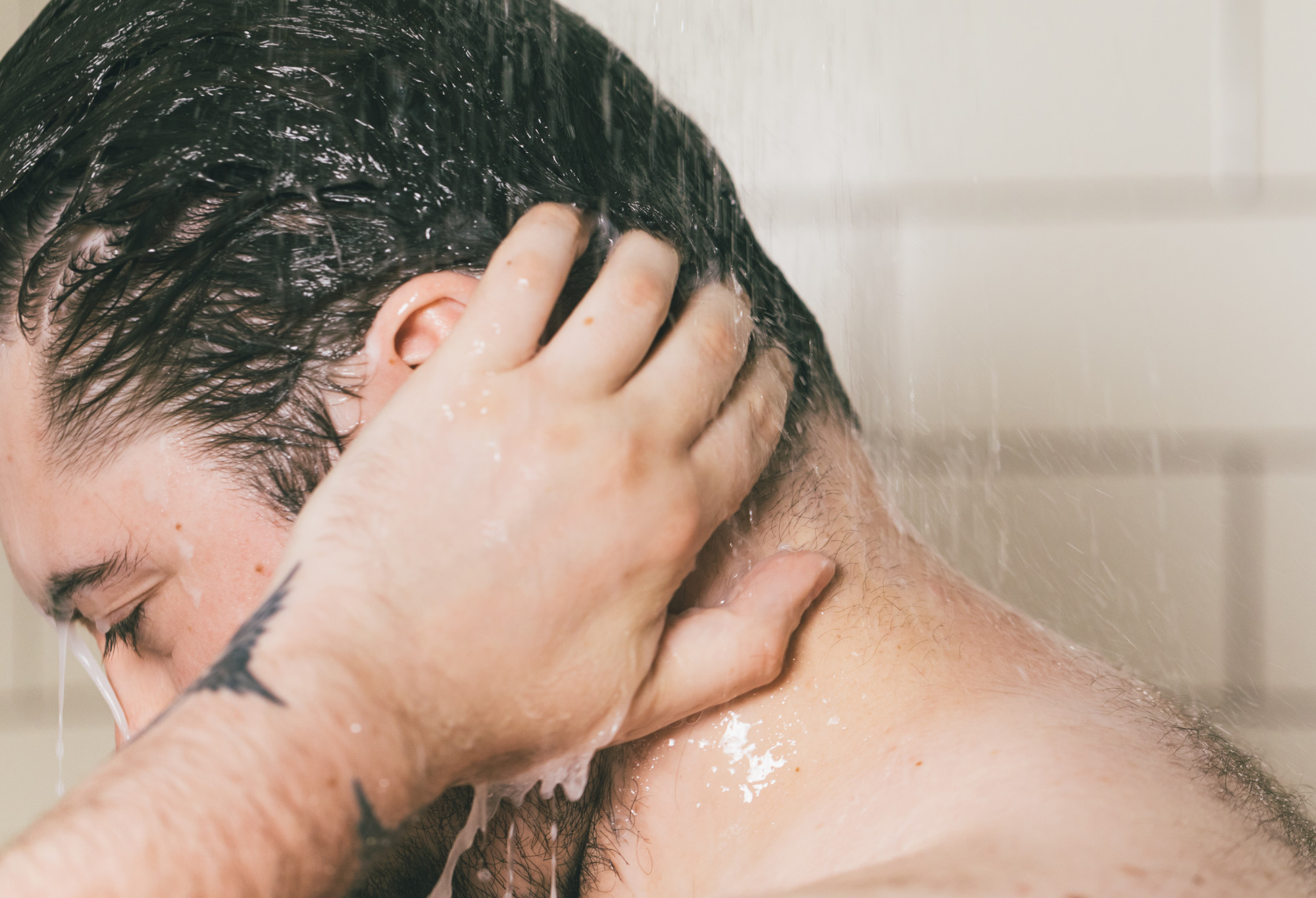 A person with short, dark hair and tatoos rinses conditioner out of their hair, creating a milky run-off of water.
