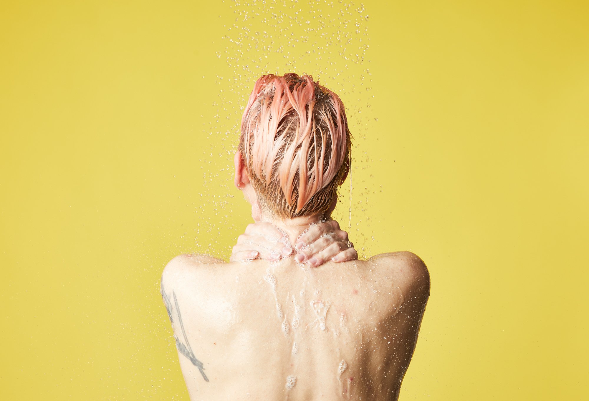 A model stands with upper back and head shown, lather running down from hands over their neck, in front of a yellow background.