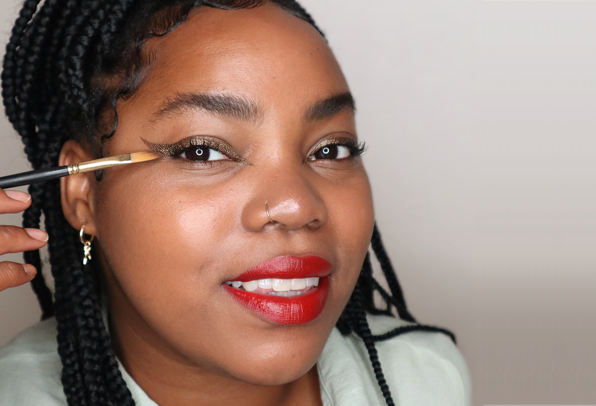 A person with braids smiles, as they apply shimmery bronze eyeliner Grounded with a Wing It Brush.