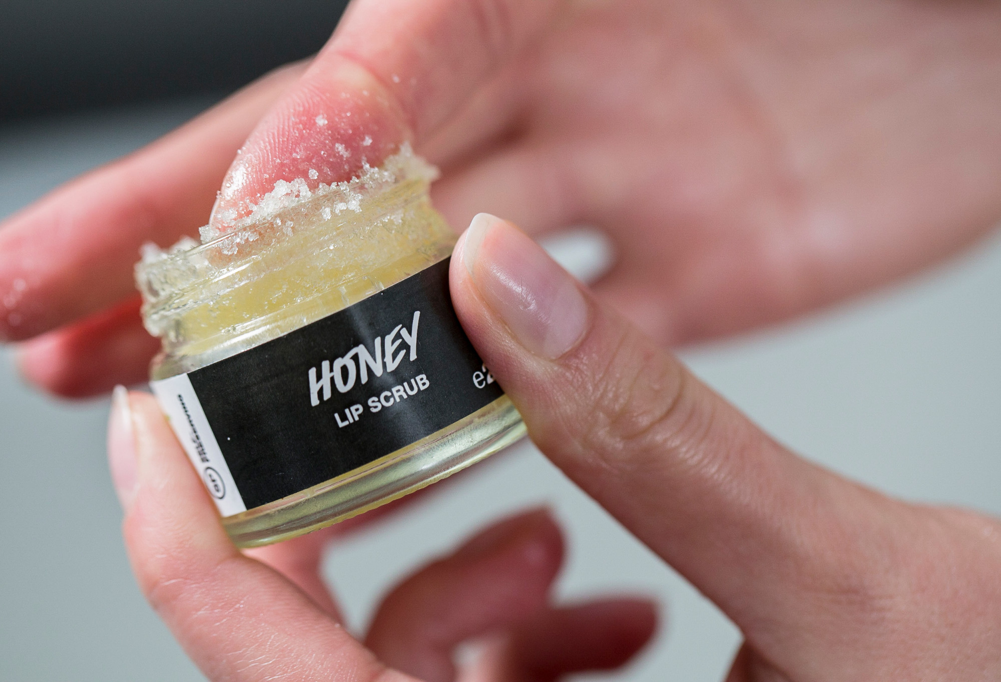 Honey, a light golden sugar lip scrub. is scooped out of its glass pot with an index finger.