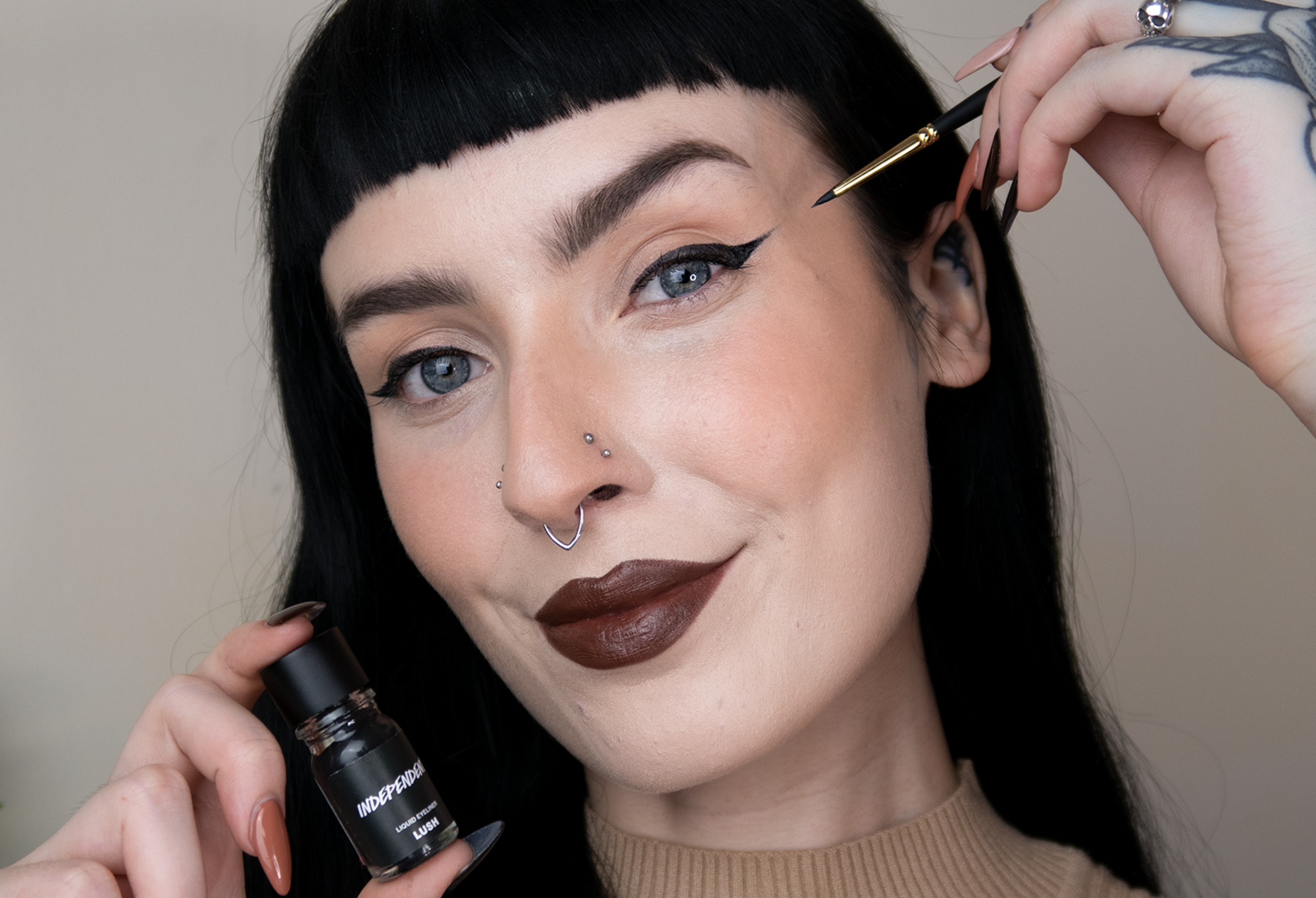 A person with dark hair and black winged eyeliner smiles, posing with a Get In There Brush and Independent eyeliner in hand.