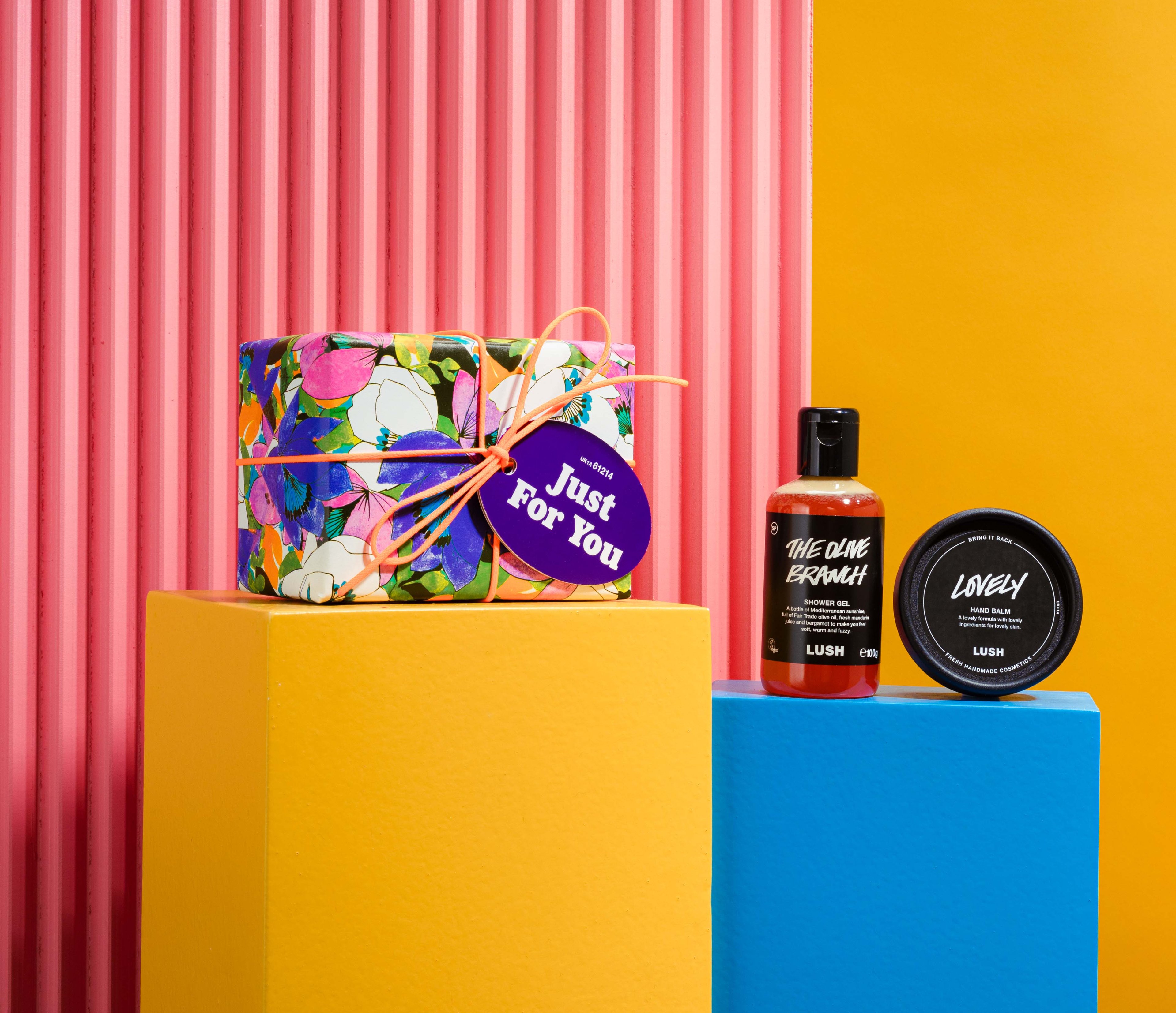 Just For You gift, in front of a pink and yellow background, surrounded by its products on blue and yellow plinths. 