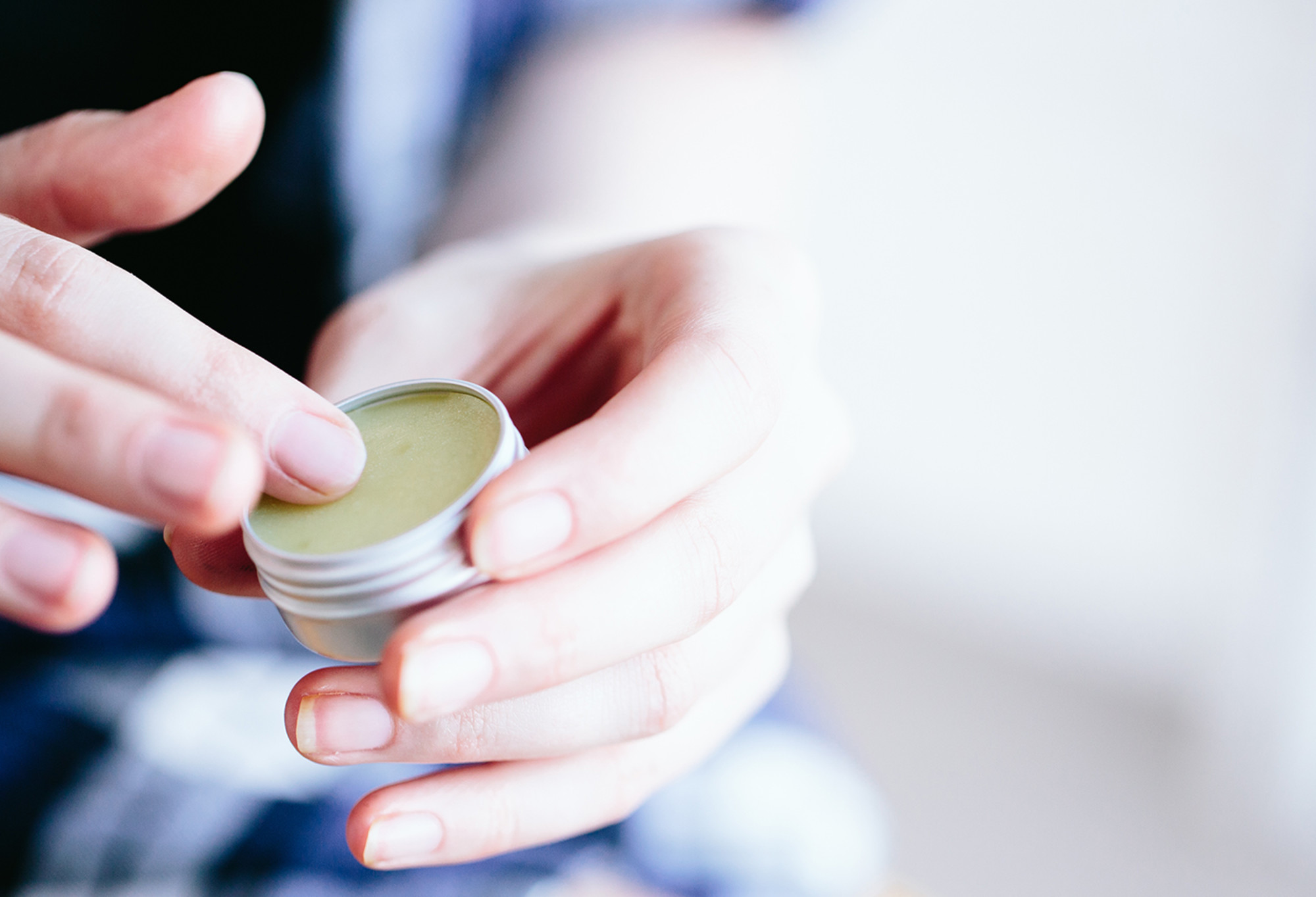 A finger is rubbed in a small round tin of thick, light green Key Lime Pie lip balm.