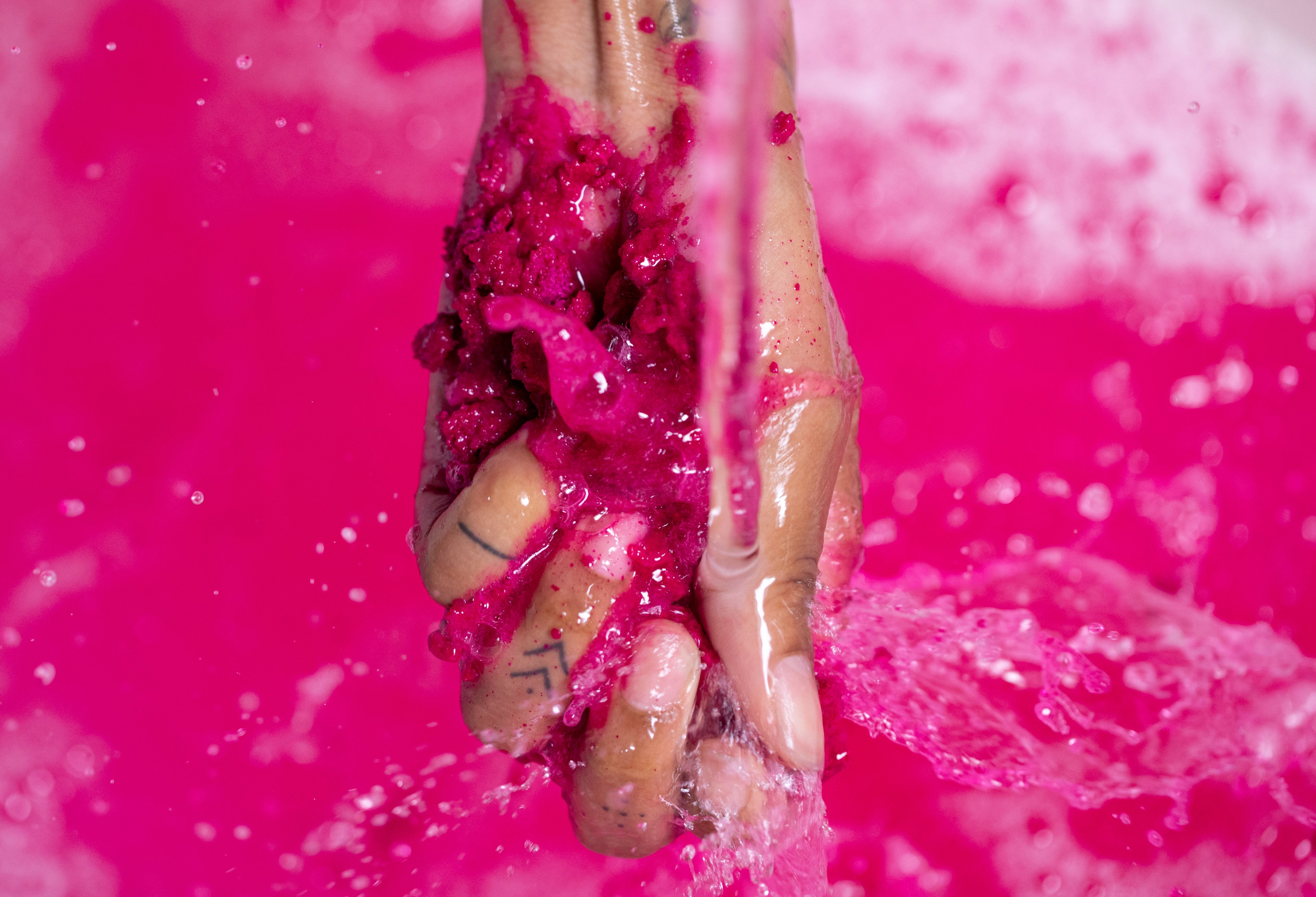 A hand crushes the bubble bar under running water, vivid pink water can be seen in the background.