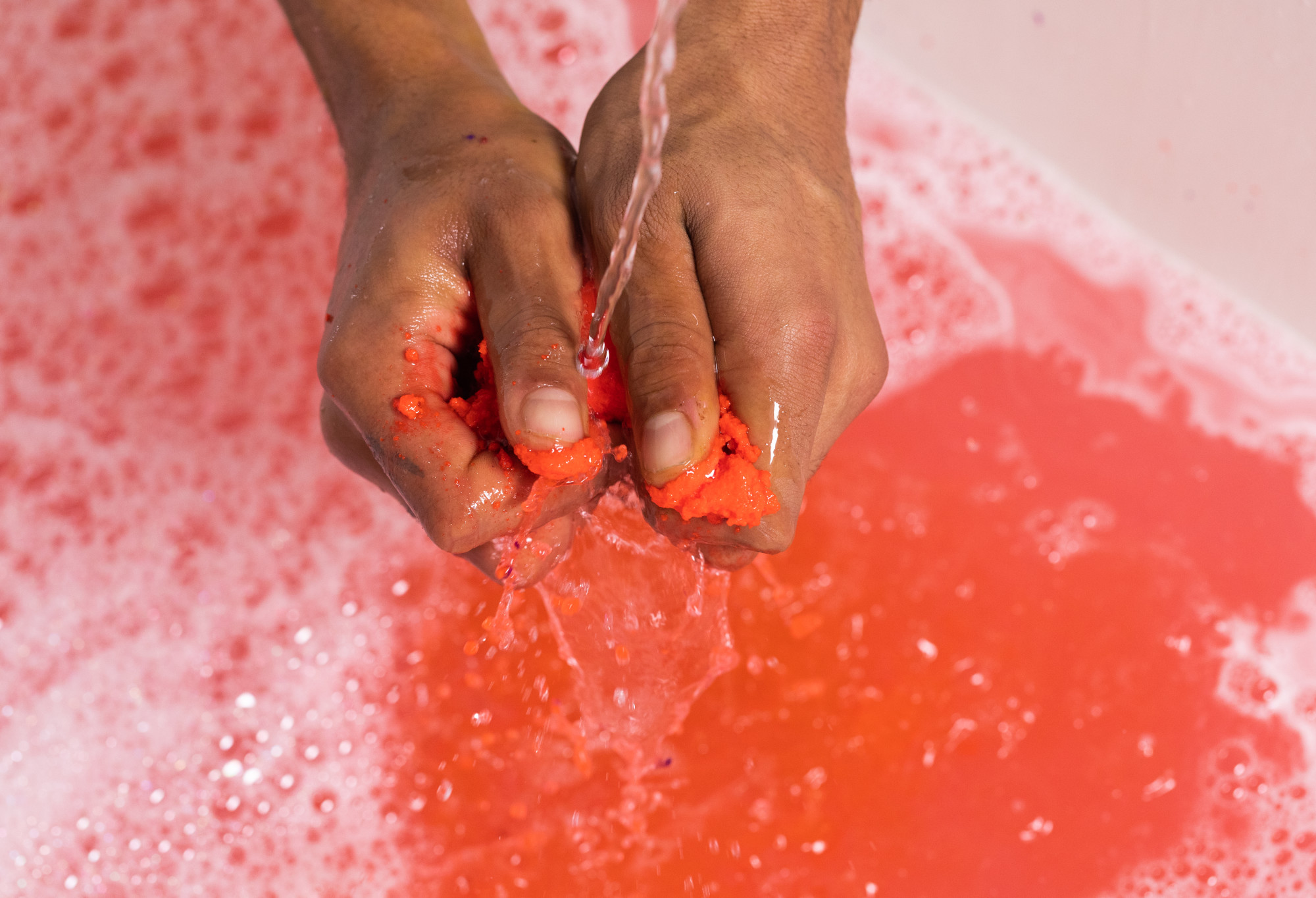 Two hands crumbling a red bubble bar under a slow running tap, adding to bubbles in a bath filled with bright, pinky red water.