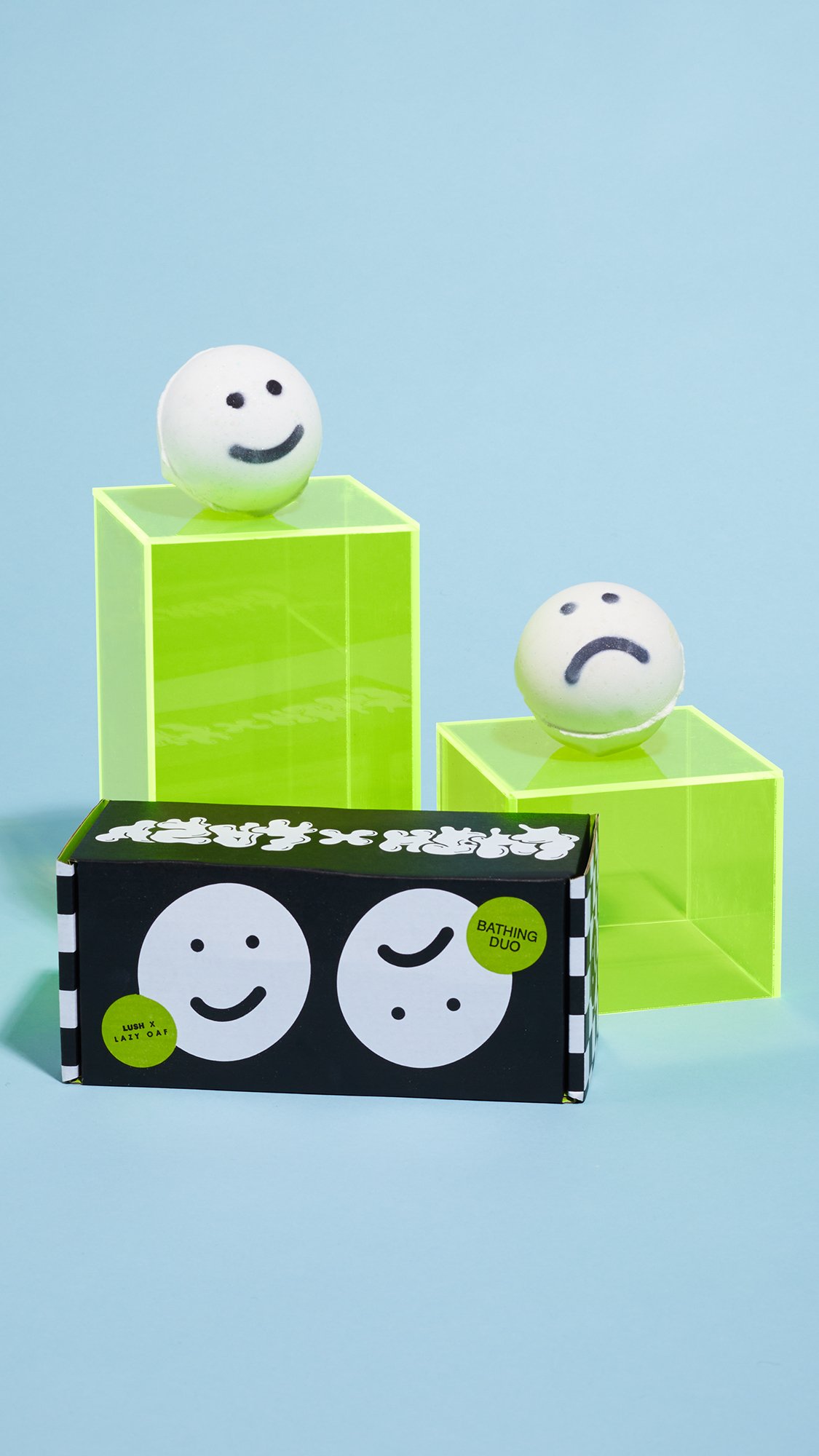 Happy Face and Sad Face sit upon green boxes, above a black, white and green packaging box. In front of a light blue background.
