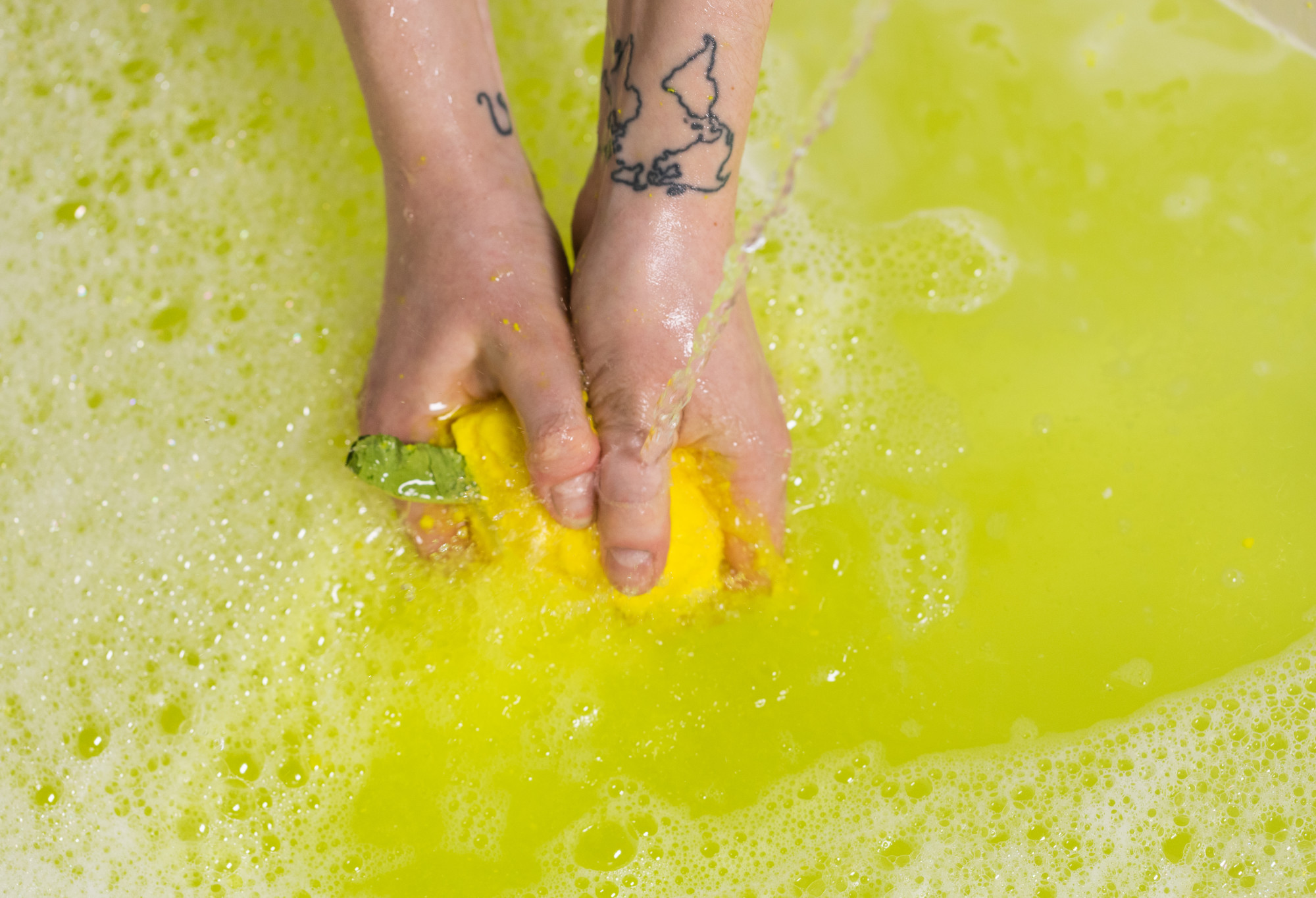 Hands crumbling the yellow Lemon Crumble in water as running water flows over. The water is bright yellow with bubbles forming.