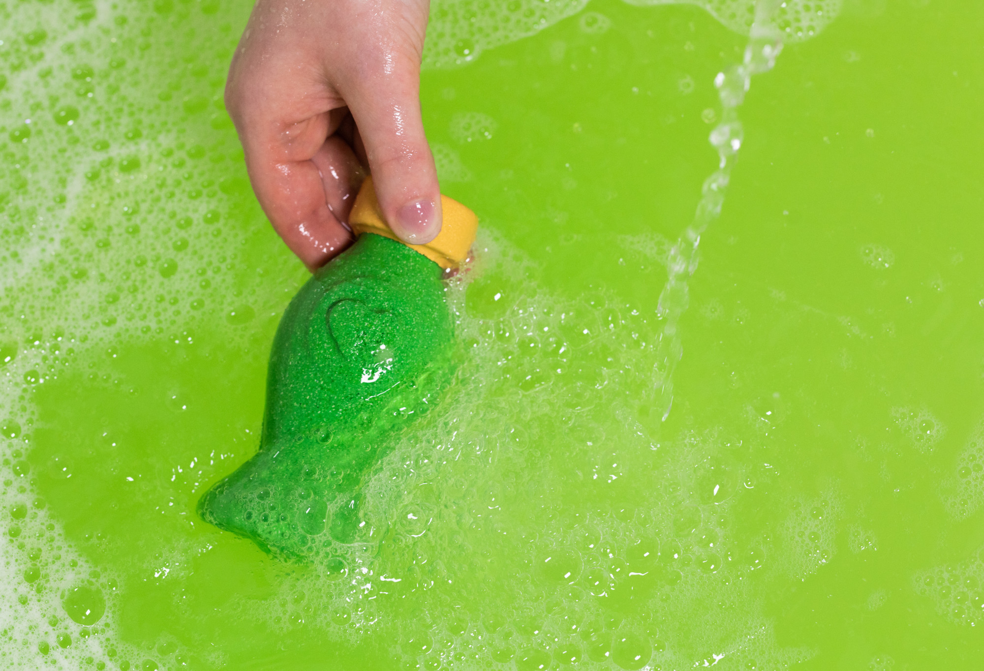 A hand is holding the golden lid of the green Love perfume bubble bottle in green water. Water is running nearby with bubbles.