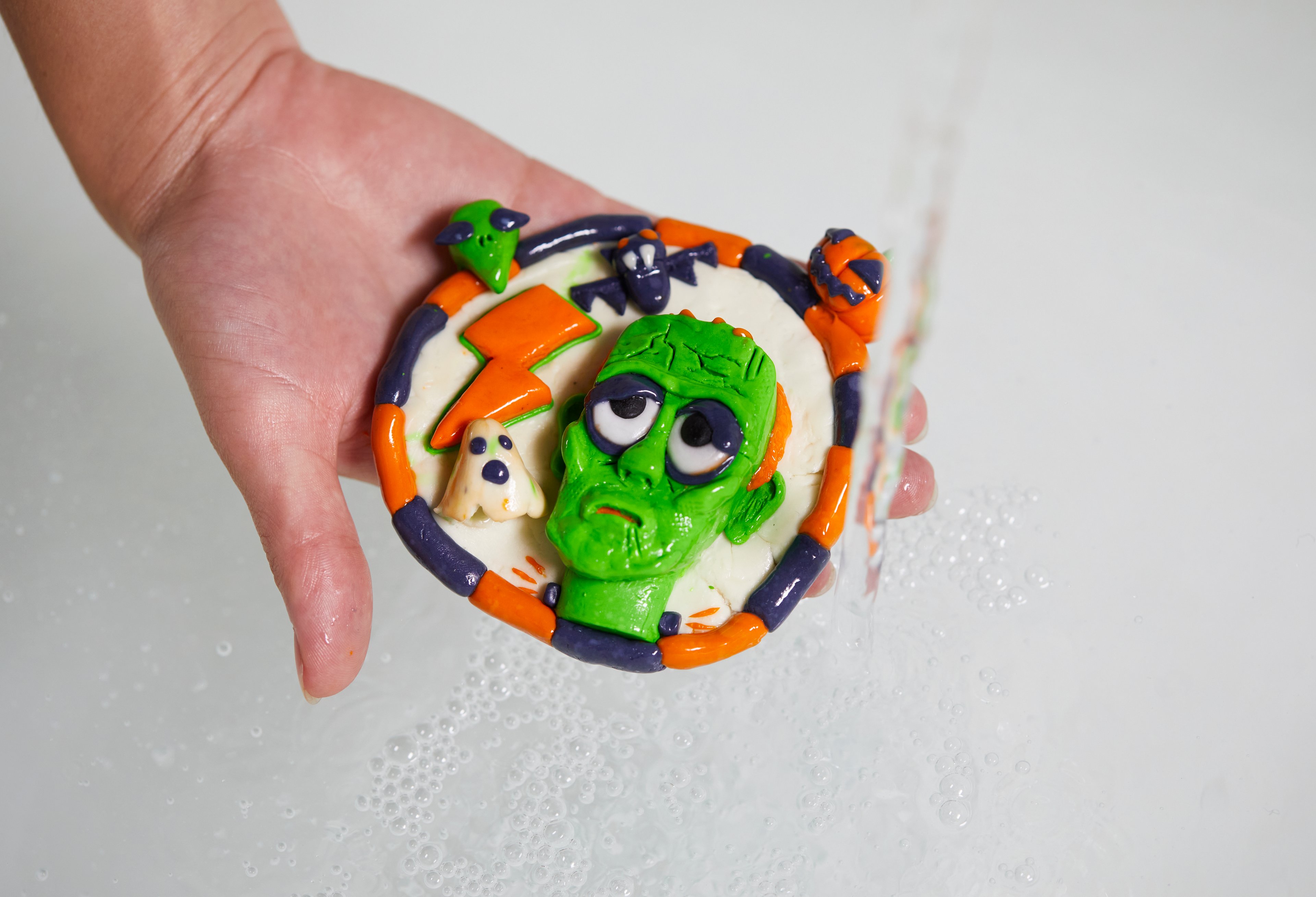 A Fun sculpted model of a green Frankenstein with orange, white and black details is held under running water.