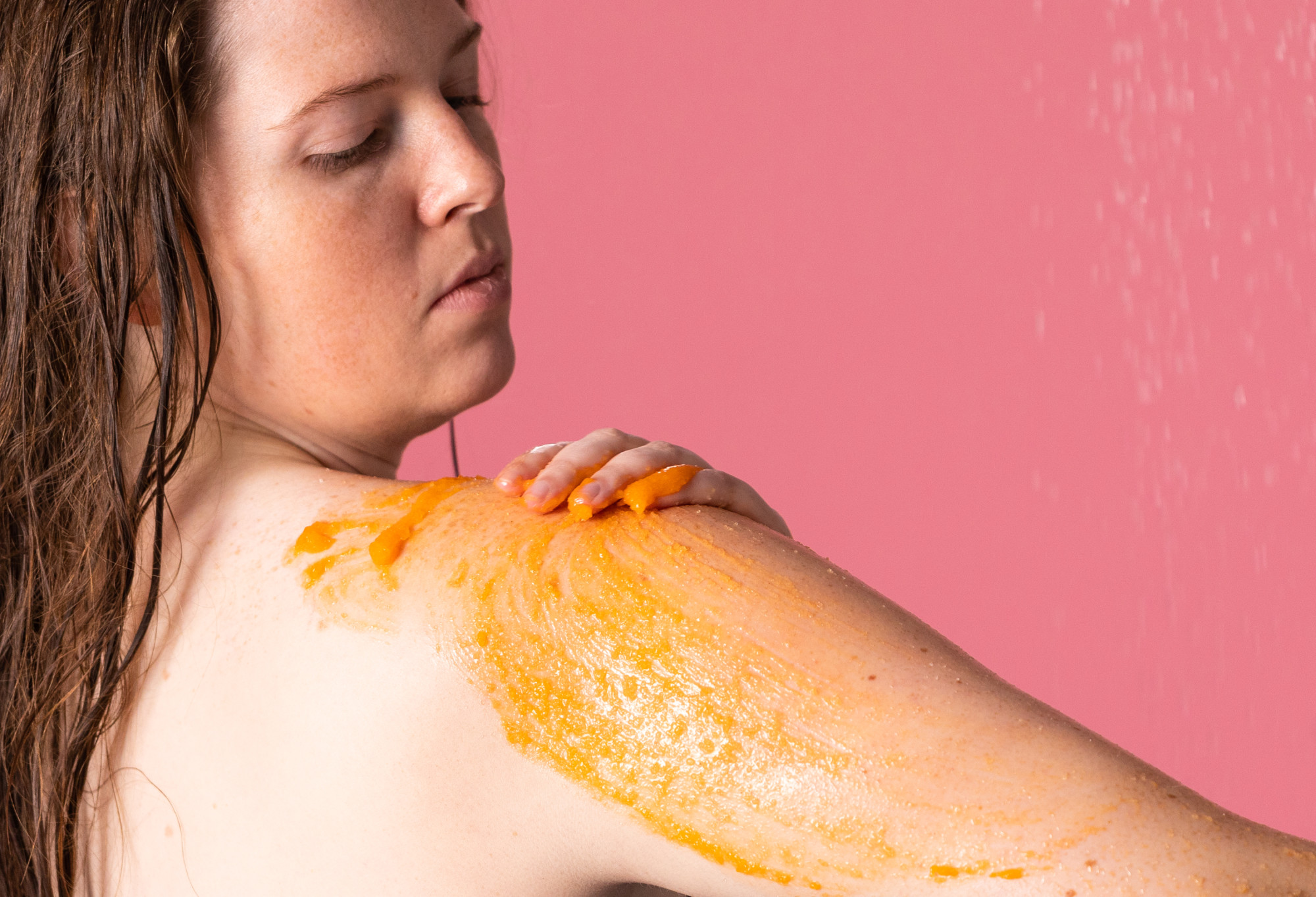 A person with long, wet hair looks down as they rub their arm, which is coated in a bright orange, sea salt body scrub.
