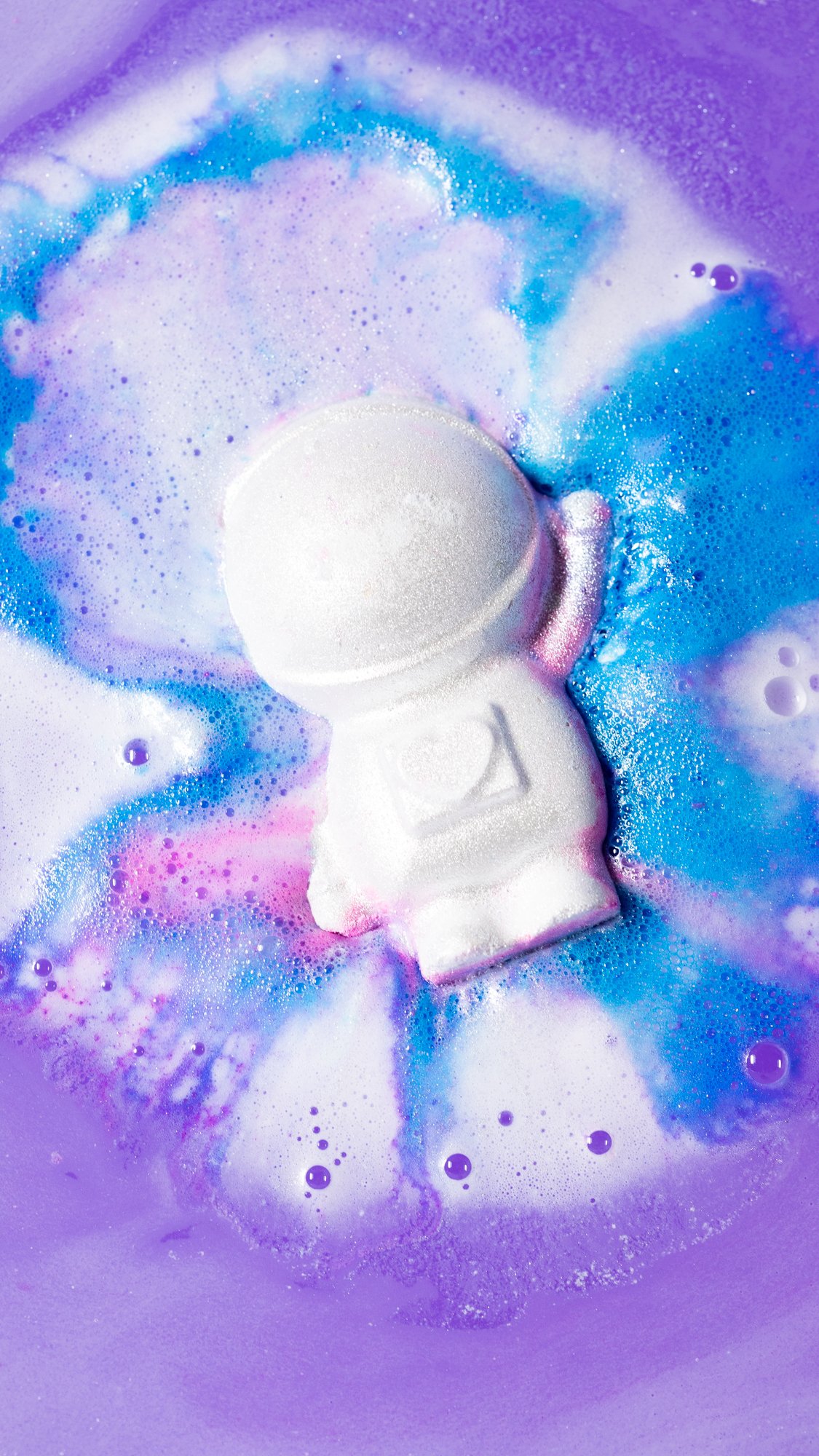 The Bath Bomb sits in the bath surrounded by foamy swirls of pink, purple and blue.