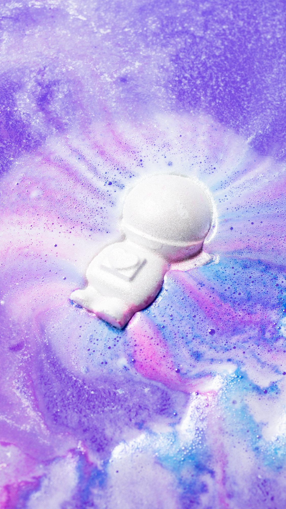 The Bath Bomb sits in the bath surrounded by foamy swirls of pink, purple and blue.