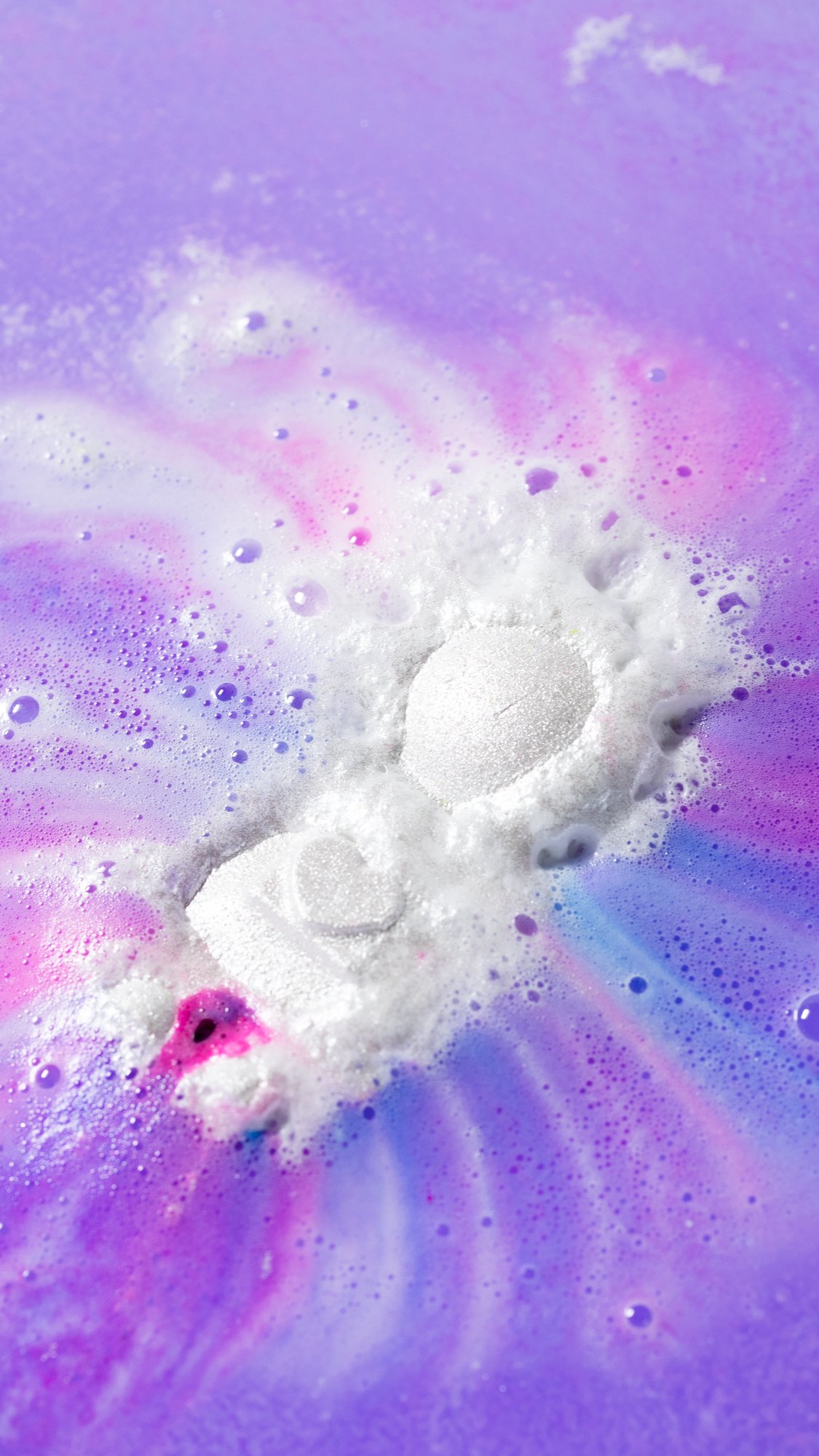 The Bath Bomb sits slightly sinking in the bath surrounded by foamy swirls of pink, purple and blue.