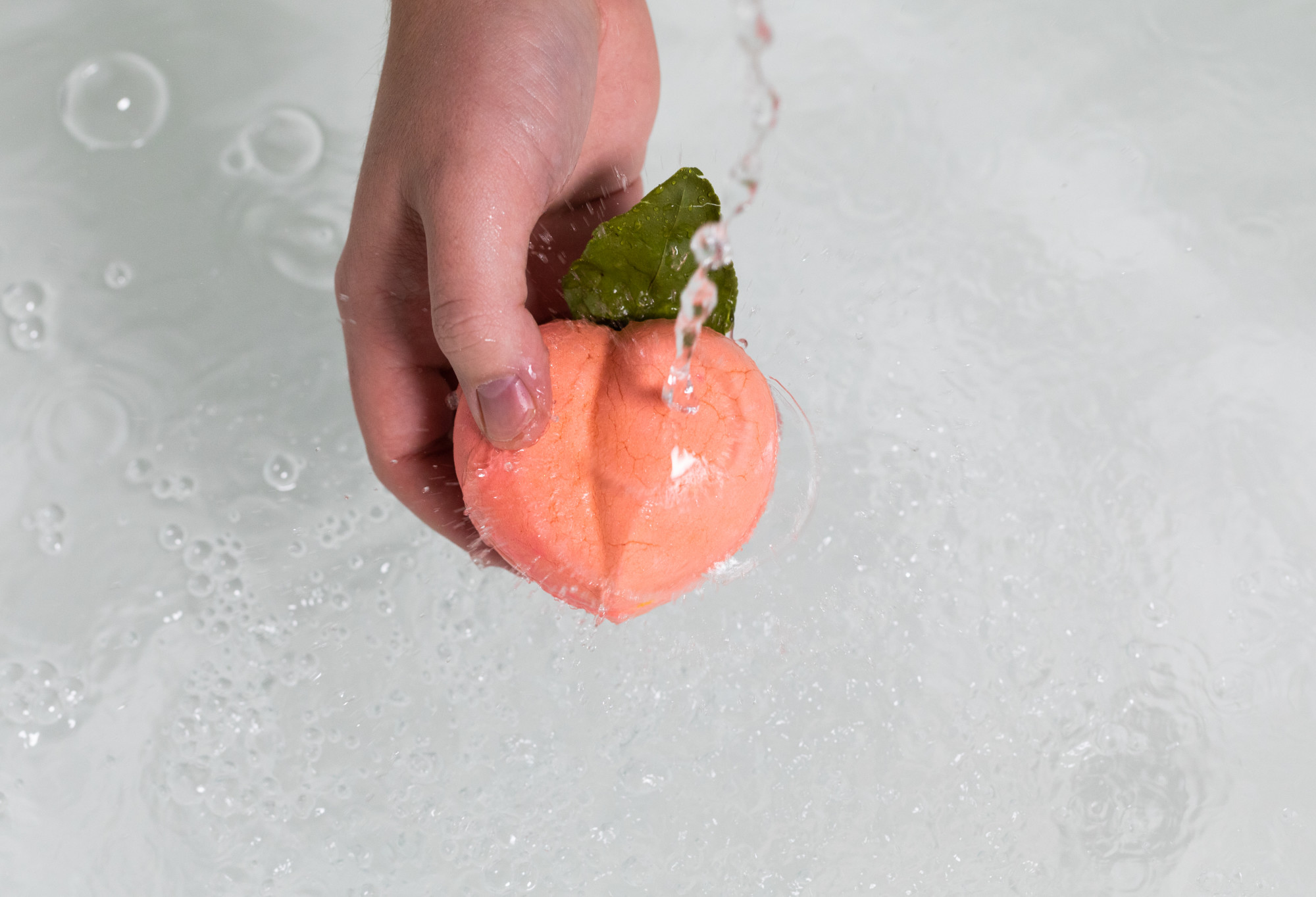 A hand holds the Peach Crumble under running water. A bubble is on the peach. In the background there's white foamy water. 