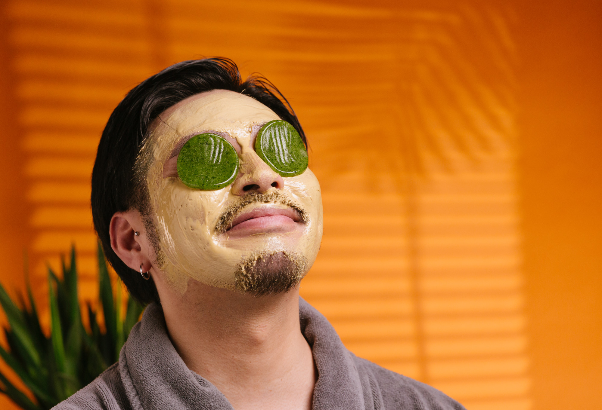 A person reclines back and smiles, wearing a gold face mask and dewy, lime green, circular Cucumber Eye Pads over their eyes.