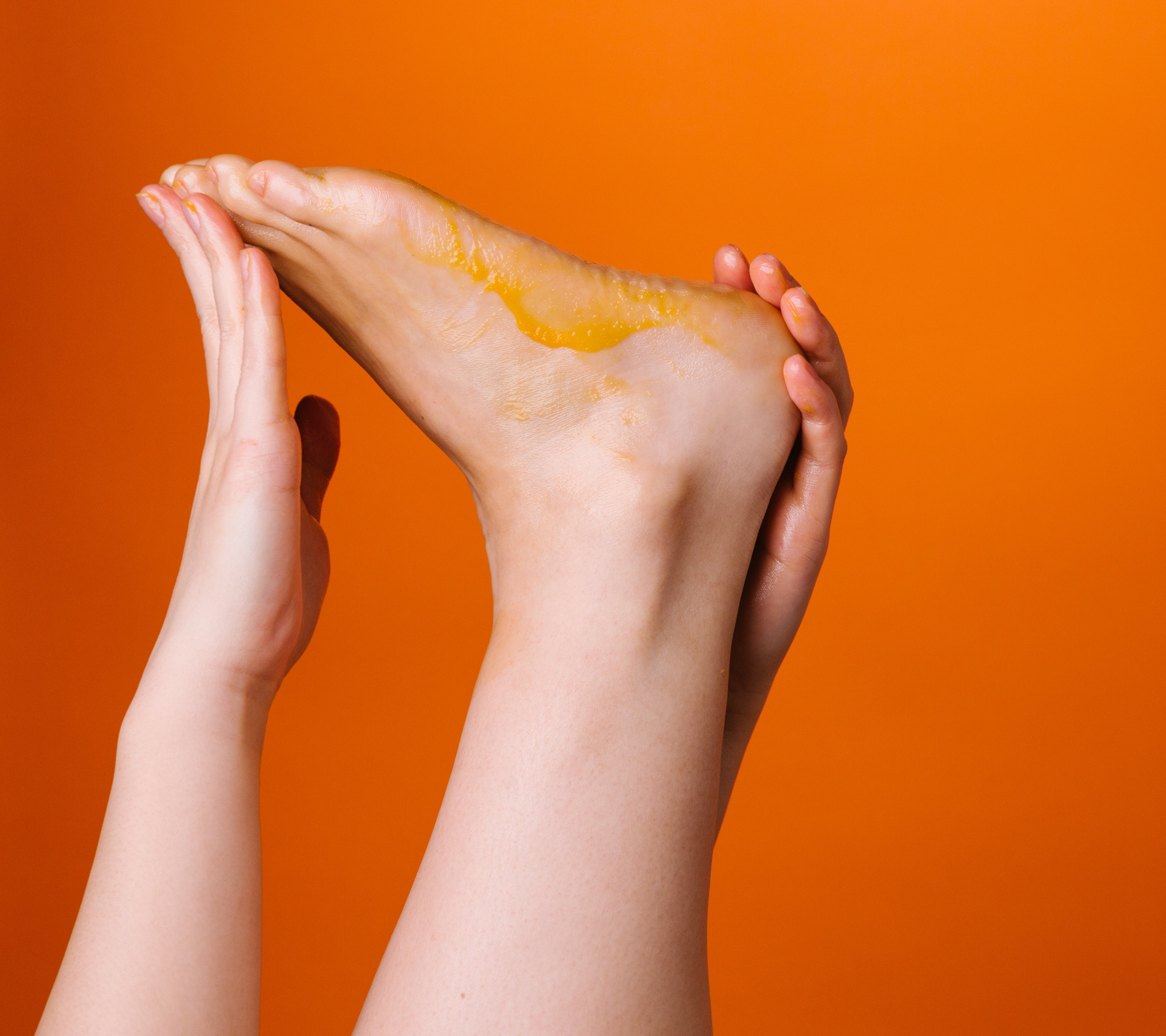 In front of an orange backdrop, a foot is being stretched by two hands, with its sole facing upwards, coated in an orange balm.