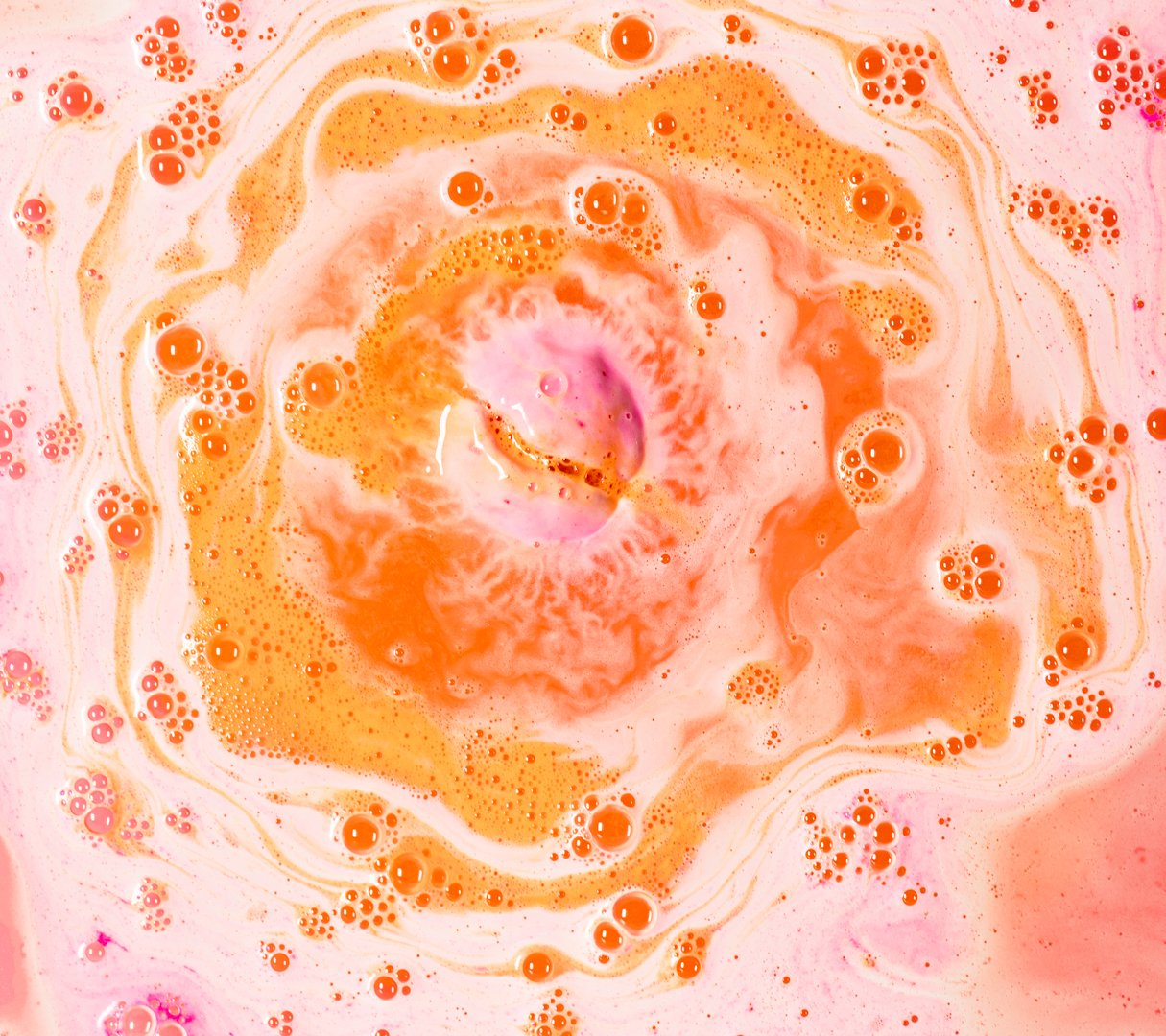 A mosaic of white and orange bubbles circle around the fizzing bath bomb, which floats in rose coloured water. 