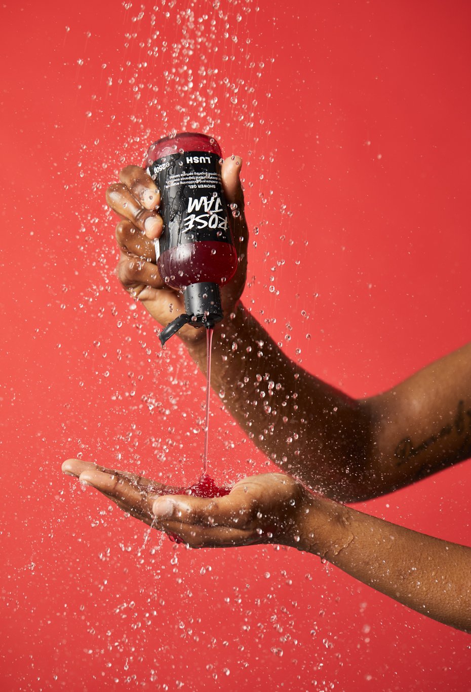 Rose Jam shower gel is being poured directly from bottle to hand, under running shower water on a deep red background.