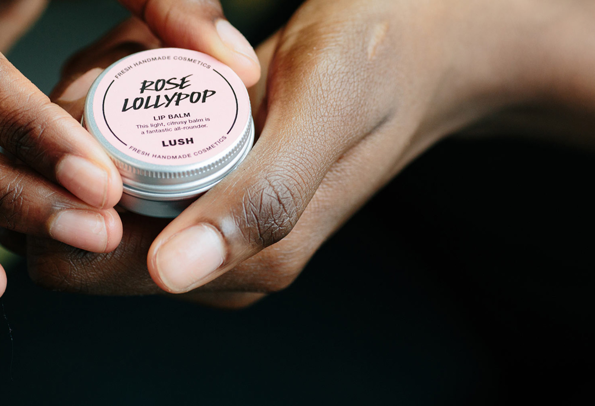 A small, metal tin, with a rosy pink label stating Rose Lollipop lip balm is held, as if about to be opened.