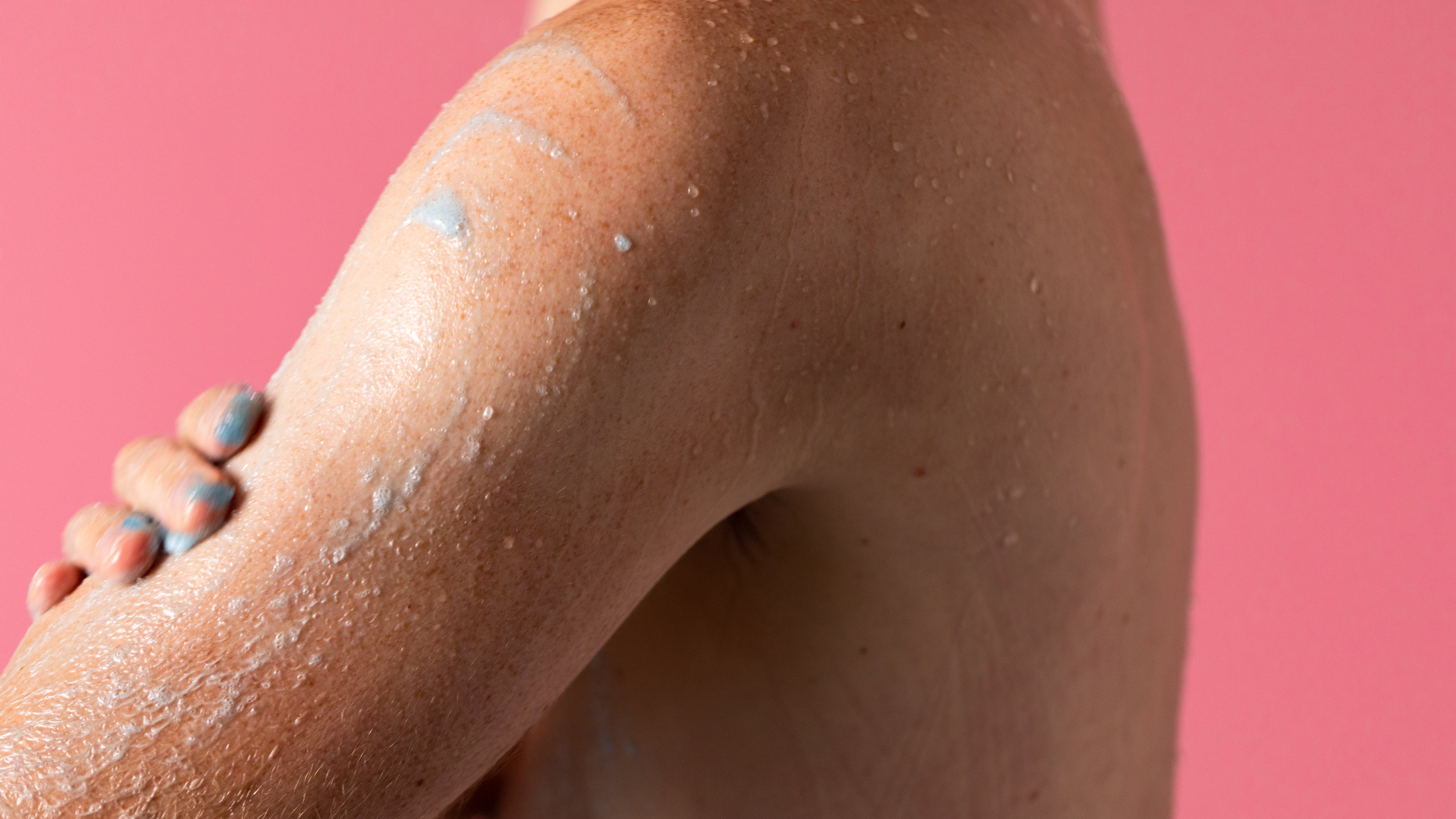 Model is using Rub Rub Rub shower scrub, gently exfoliating the shoulder and upper arm, on a pink background.