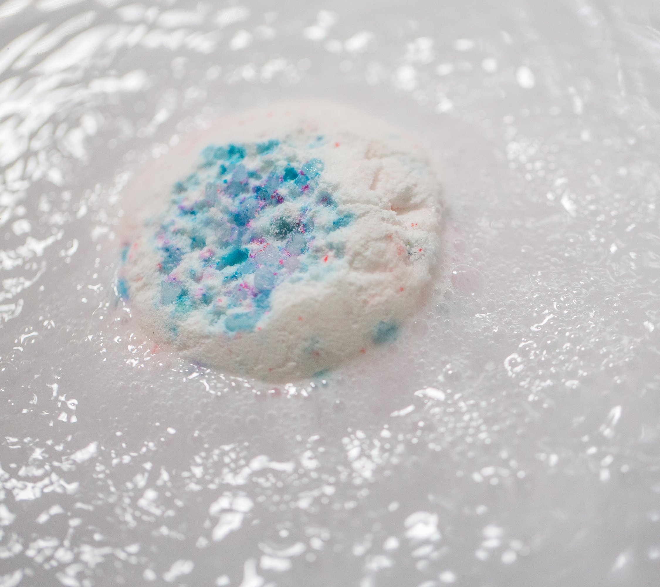 A close-up of a semi-submerged Sakura bath bomb, pink-flecked white with blue-coloured salt, fizzing in milky white waters.