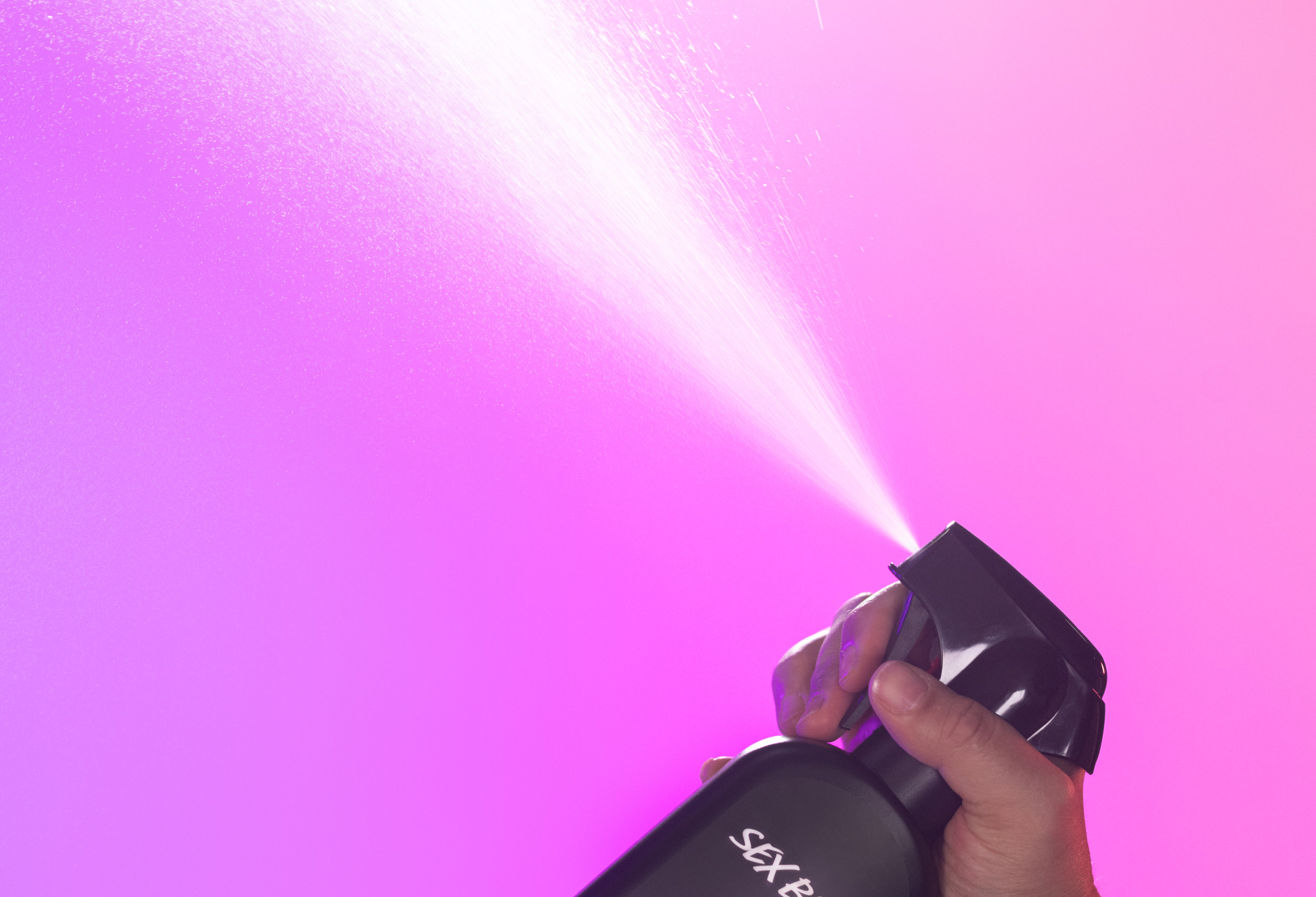 Sex Bomb body spray is sprayed up into the air, in front of a vibrant, purple to pink gradient background.
