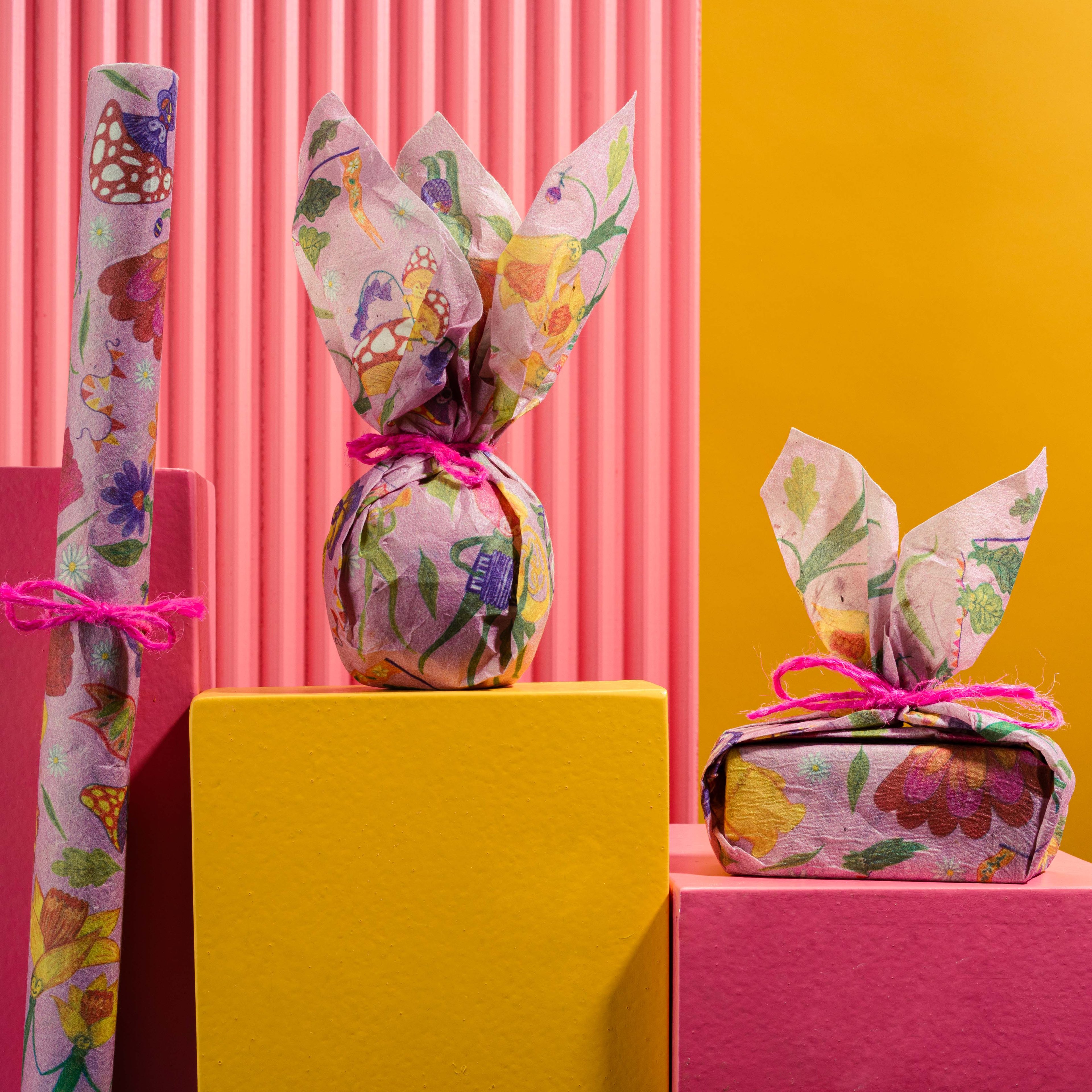 The Lokta Wrap is decoratively wrapped with a pink ribbon, alongside one folded, in front of a pink and yellow background. 