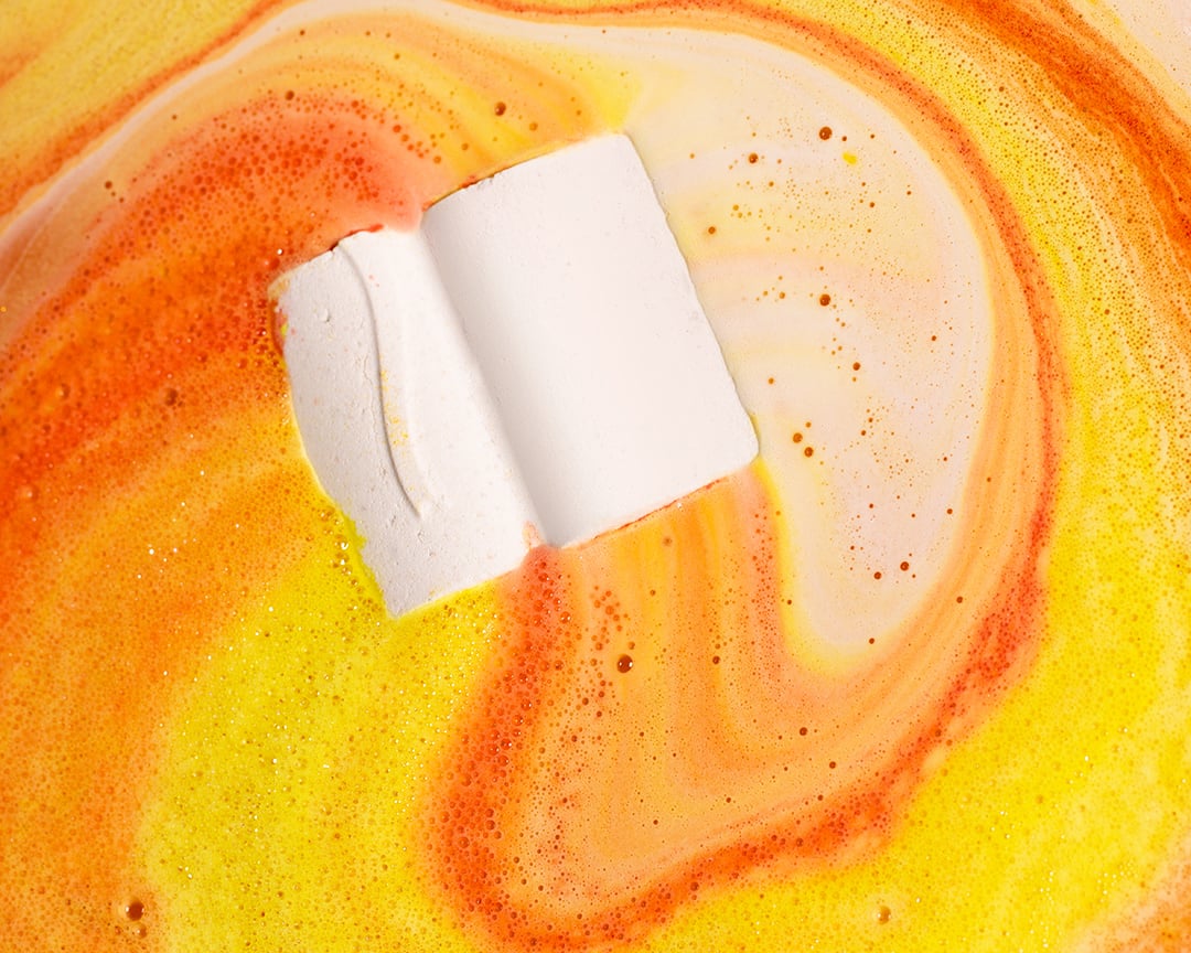 The book-shaped of Knowledge bath bomb floats on the water surrounded by a spiral of orange and yellow bath art.