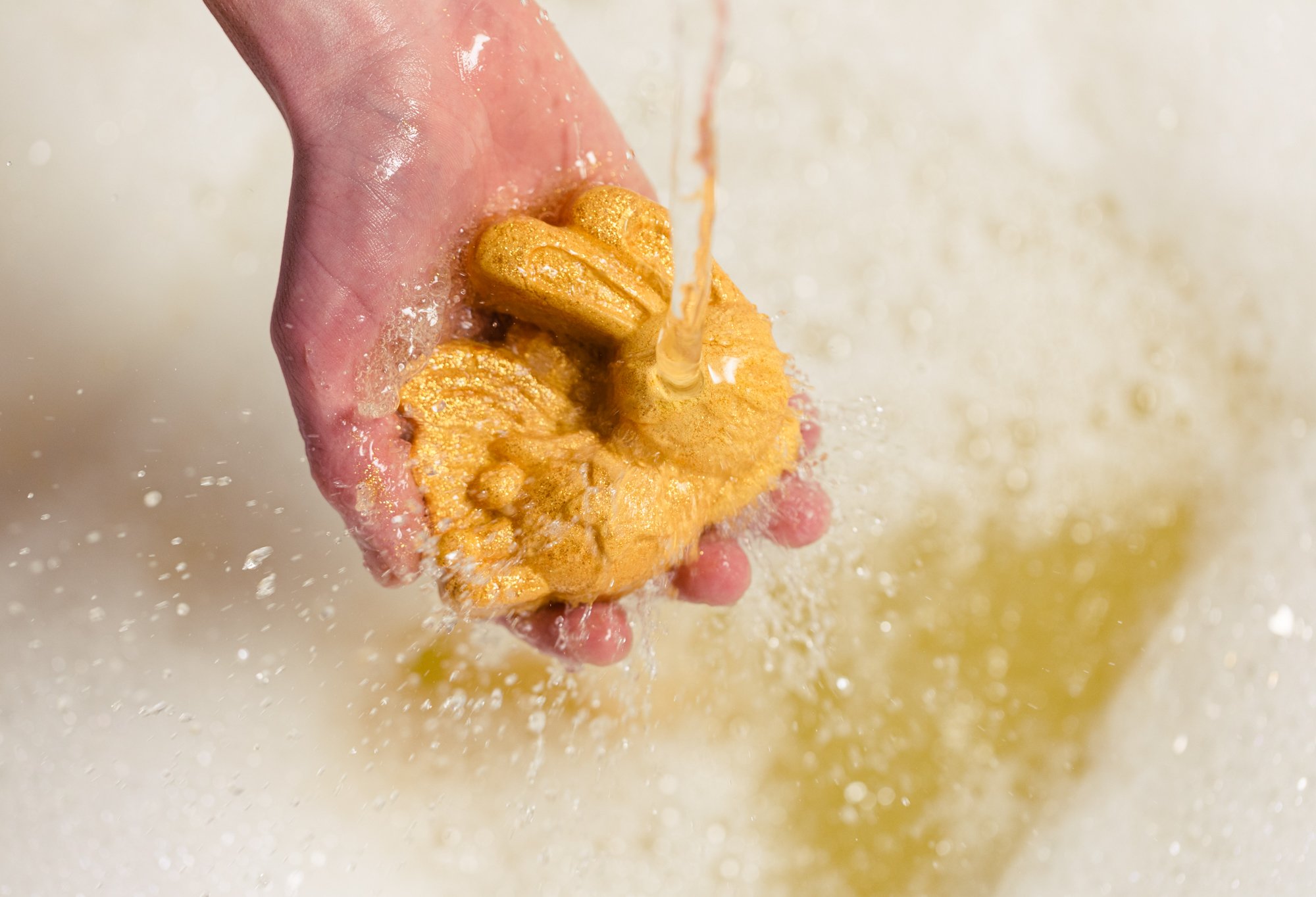 The golden bubble bar is held whole under running water, creating thick white bubbles and golden yellow water. 