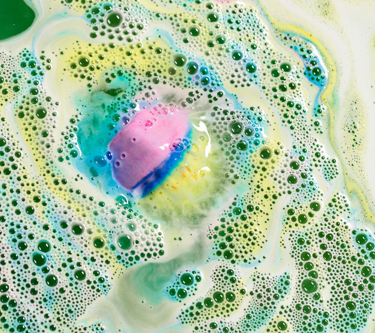 A pink, blue and yellow coloured bath bromb fizzes in bubbly waves of pastel yellow and blue foam, revealing green water below.