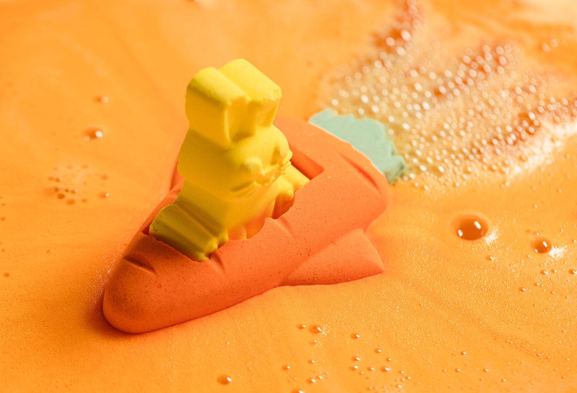 The carrot bath bomb floats in orange foamy water, with bubbles coming out its green end, the bunny bath bomb pilot sits inside.