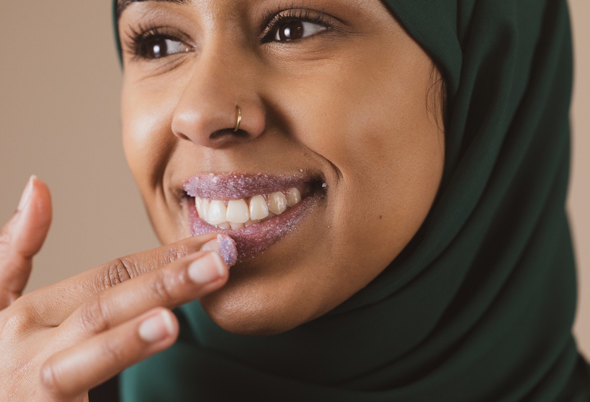  A person with a nose piercing, wearing a Hijab, beams as they scrub their lilac, Unicorn sugar lip scrub coated lips.