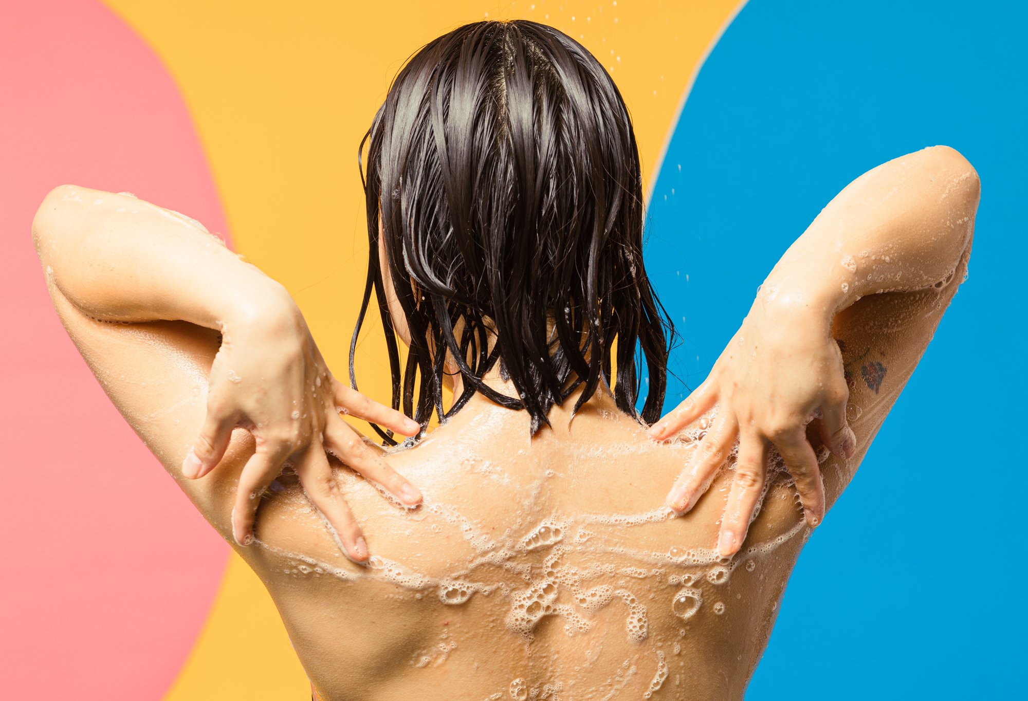 A model is shown massaging shower gel across their back and shoulders, creating light white foam and bubbles.