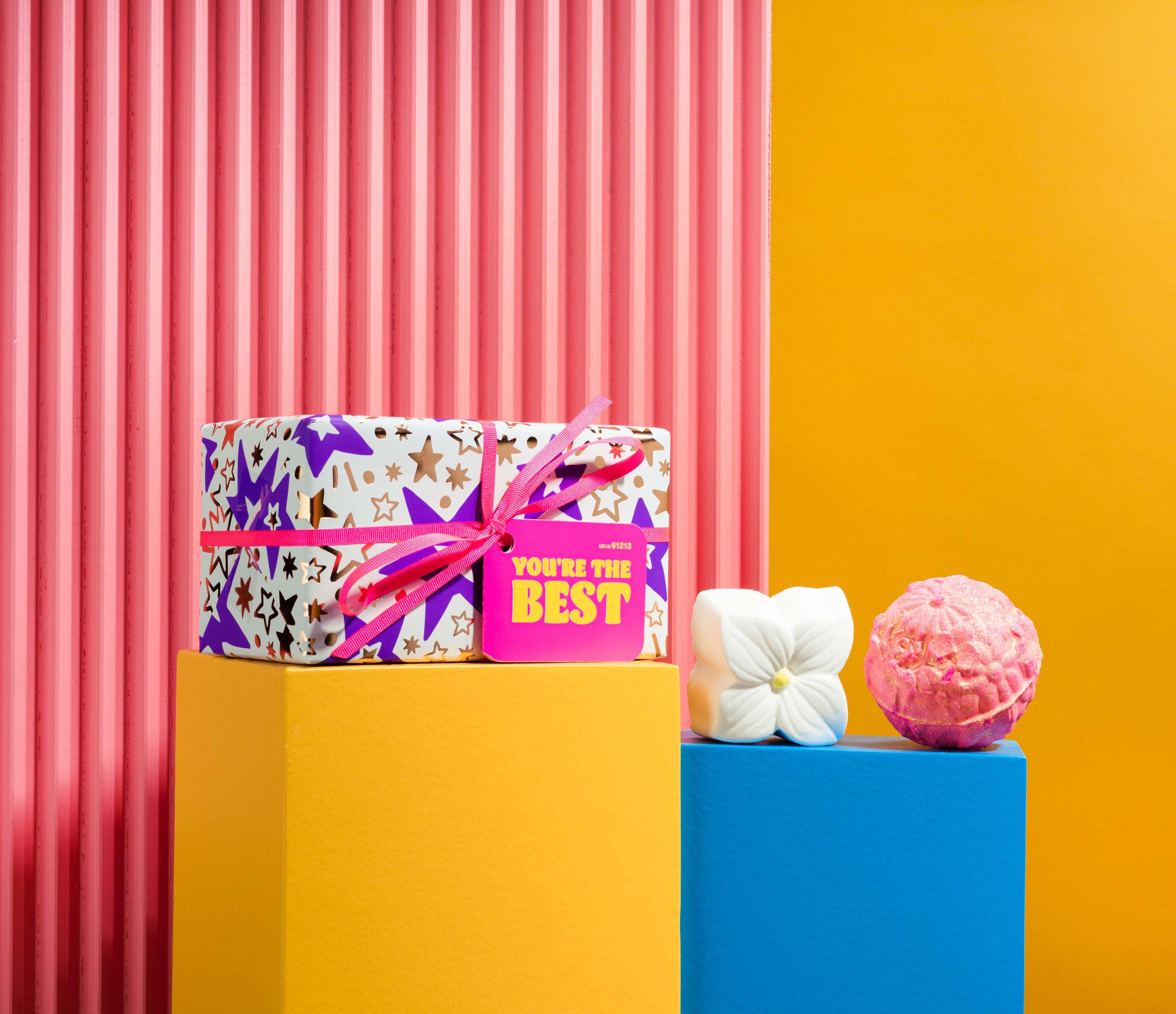 You're The Best, in front of a pink and yellow background, surrounded by its products on blue and yellow plinths. 