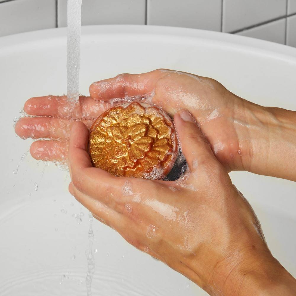 Image shows a close-up as the model washes their hand with the golden Cempasúchil soap under running water.