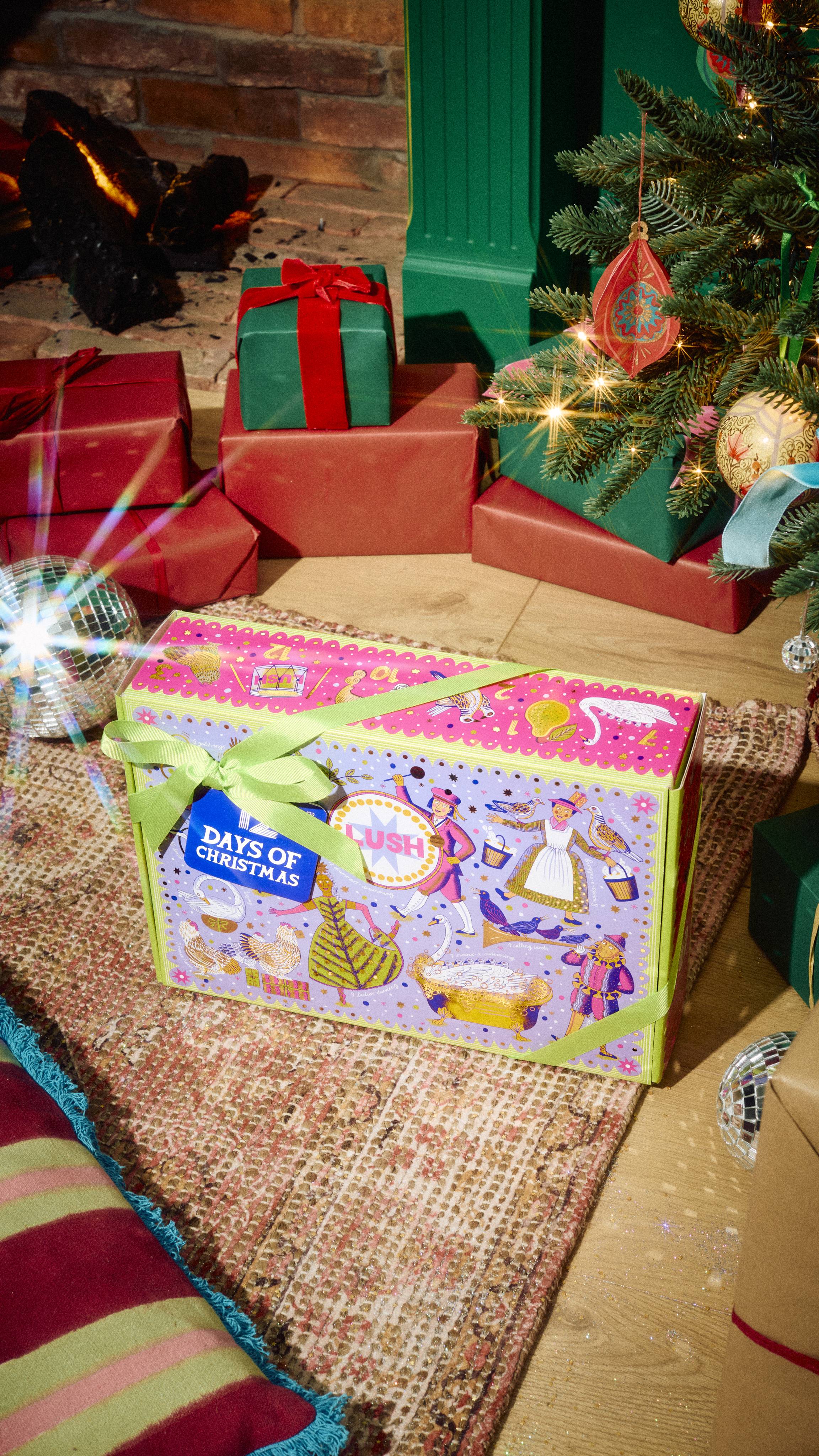 The closed Lush 12 Days of Christmas gift box, decorated with festive illustrations and tied with a green ribbon, sits on a patterned rug near wrapped presents and a decorated Christmas tree.