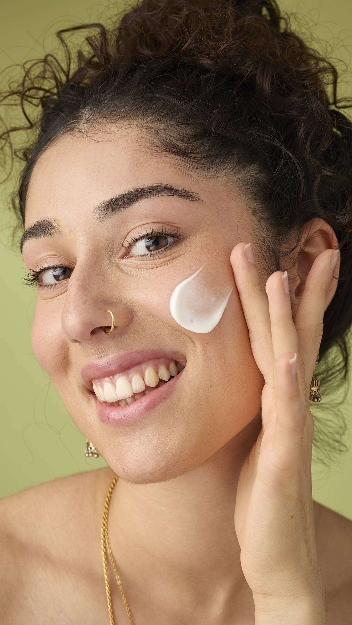 A person with dark, curly hair tied up and a nose ring smiles radiantly while gently applying the creamy white 9 to 5 cleanser to their cheek with their fingertips, enjoying the product's texture