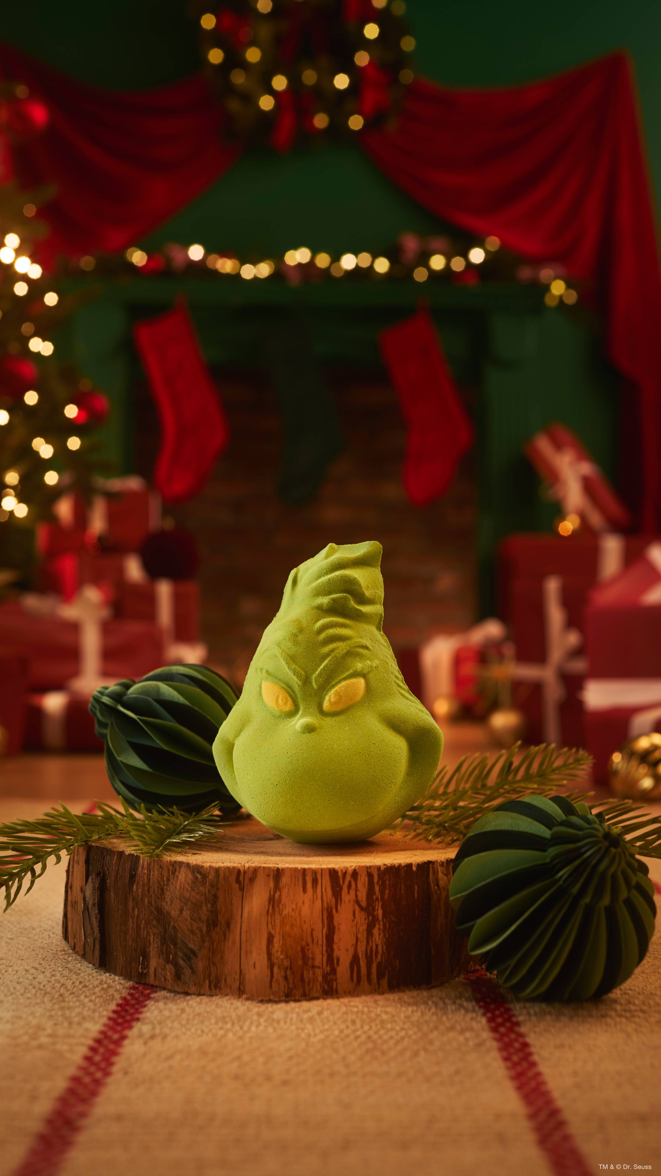 A lime-green Lush bath bomb shaped as a mischievous "Grinch" face sits on a wooden log slice alongside pine sprigs and dark green paper baubles, set against a blurred Christmas scene with fairy lights and stockings.