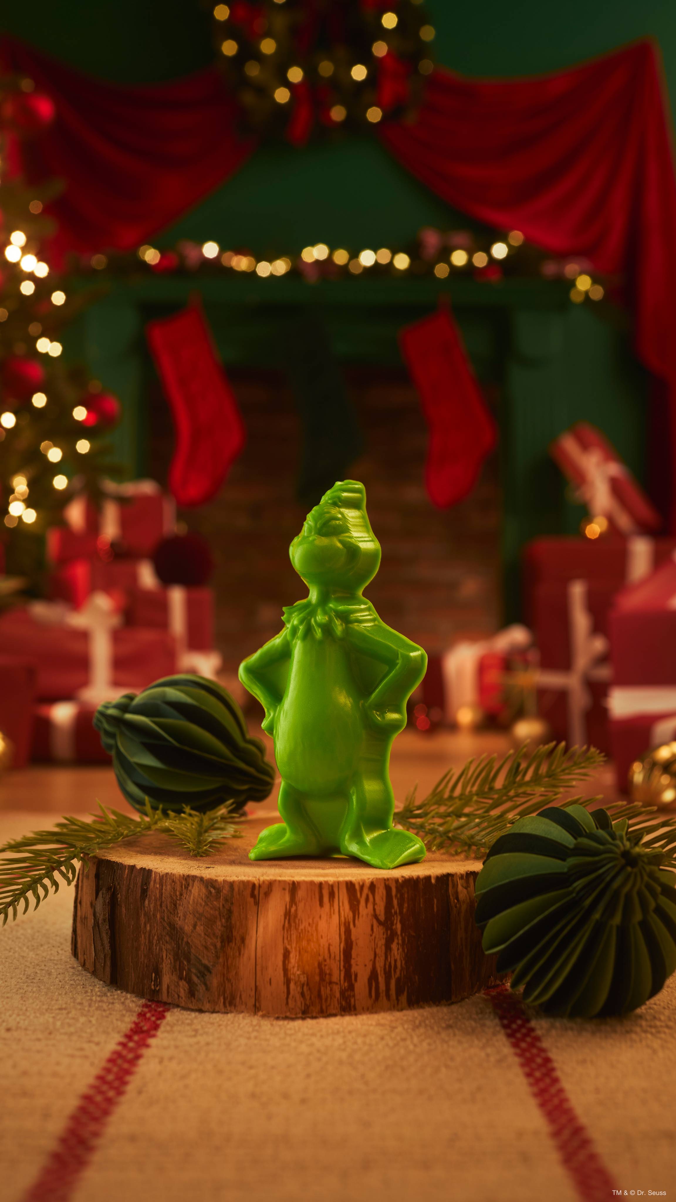 A bright green Lush shower gummy shaped like The Grinch stands on a wooden log slice beside dark green paper baubles, set against a warm, blurred Christmas scene with fairy lights and red drapes.