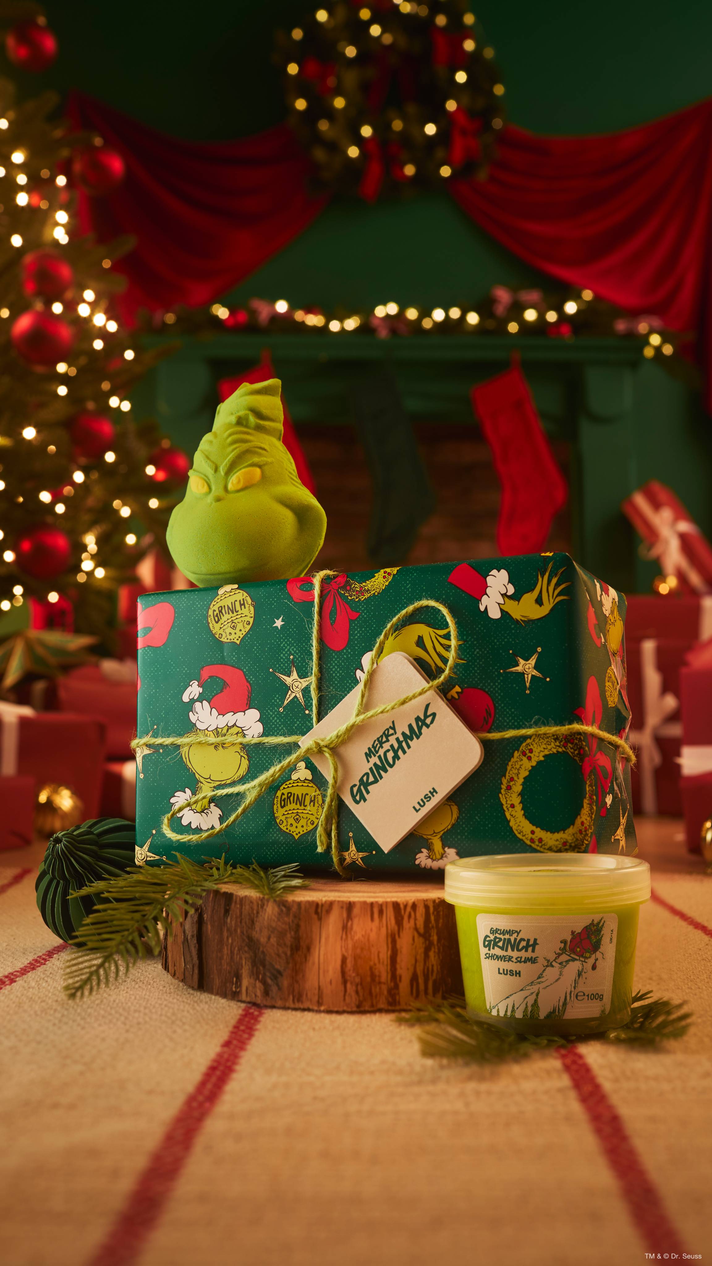 A Lush 'Merry Grinchmas' wrapped gift box sits on a log slice topped with a green character bath bomb, with a tub of shower slime beside it, set against a warm, festive Christmas background.  